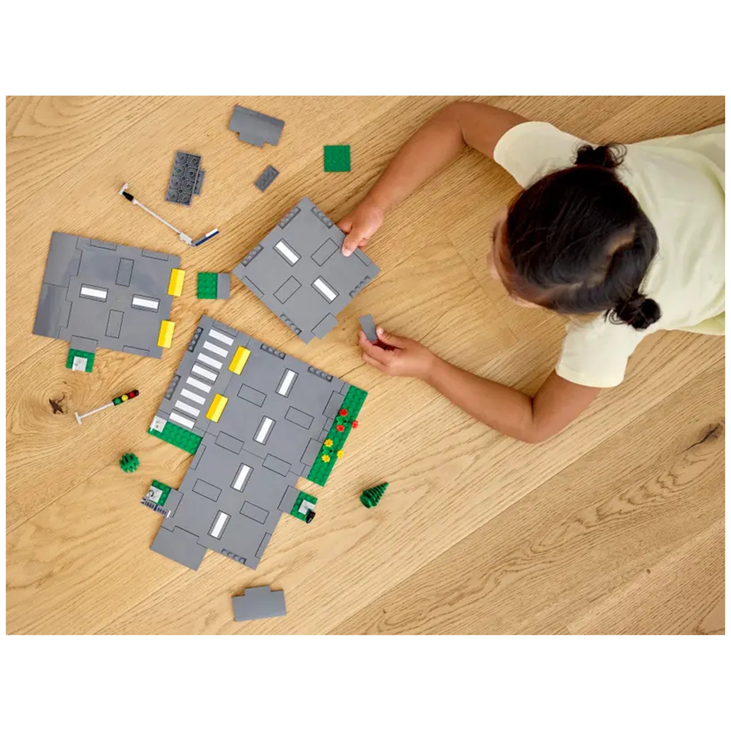 A child is assembling LEGO® City Road Plates on a wooden floor, surrounded by gray road plates, green pieces, and small accessories. The child holds a gray block, focusing on building a vibrant urban landscape.