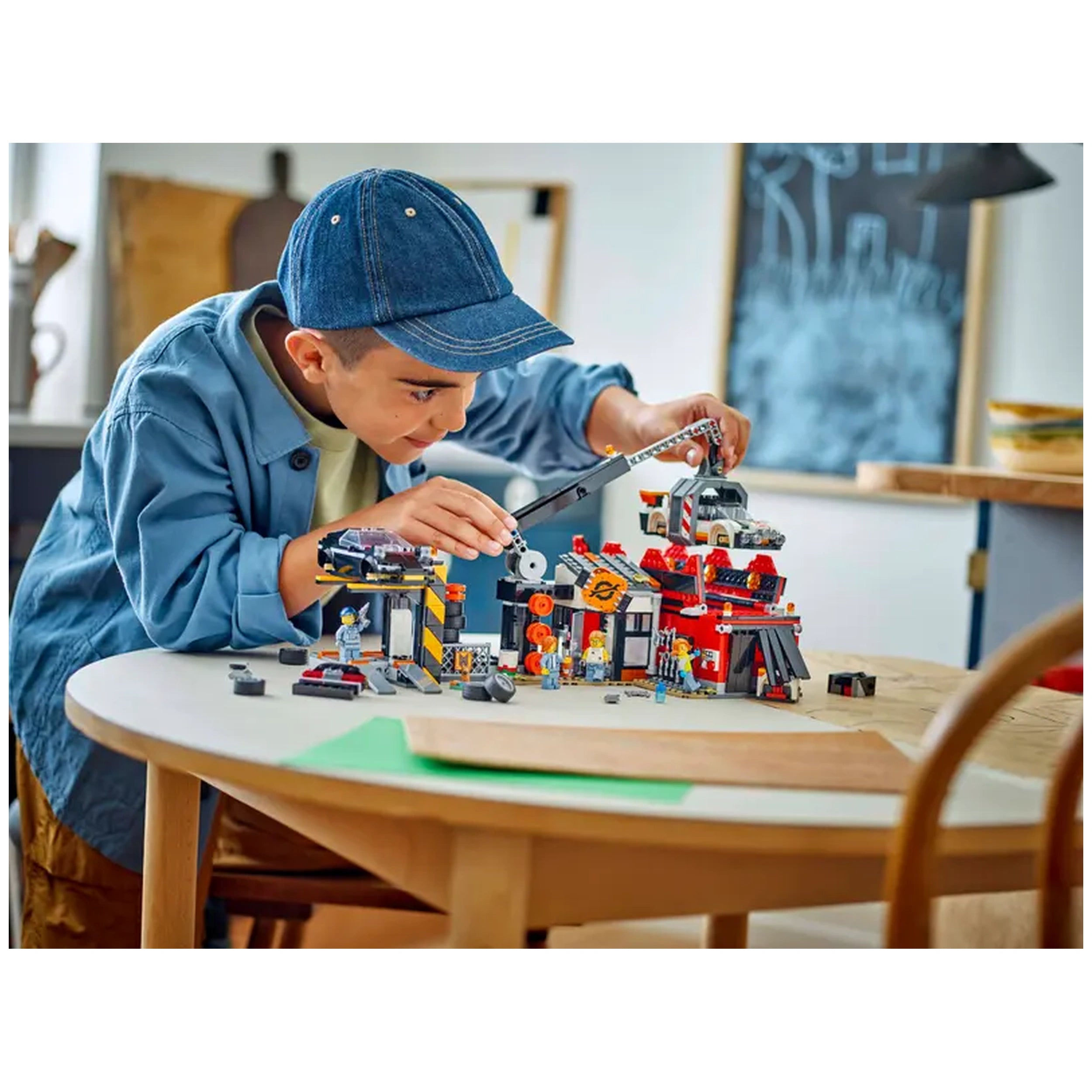 A young boy in a blue cap joyfully plays with a colorful LEGO® City Scrapyard construction set on a round wooden table, focusing intently as he uses a claw crane to lift a toy car. The vibrant playset features detailed elements like a recycling station and two dismantled classic cars, surrounded by various LEGO pieces. Natural light illuminates the serene indoor space, enhancing the playful atmosphere.