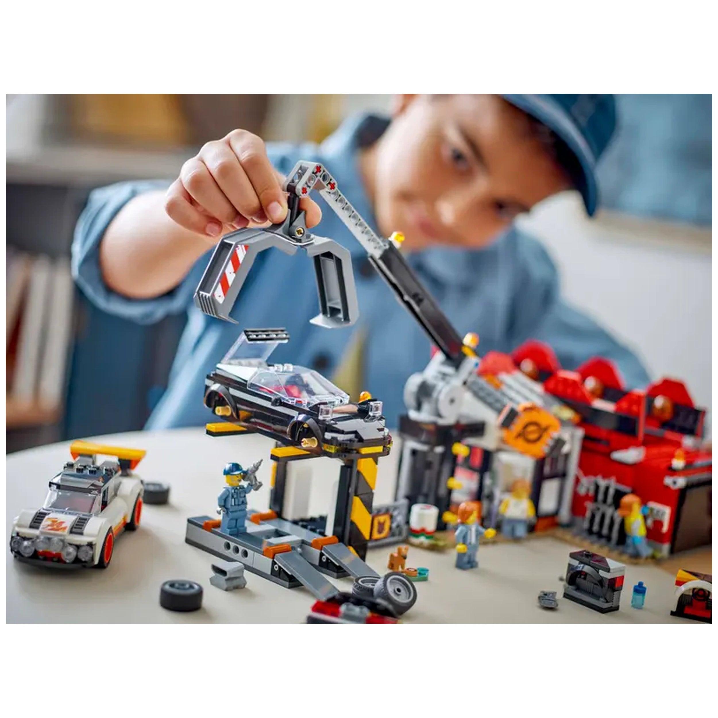 A child in a cap uses a claw crane to lift a toy car at a colorful LEGO City Scrapyard set, surrounded by various buildable elements.