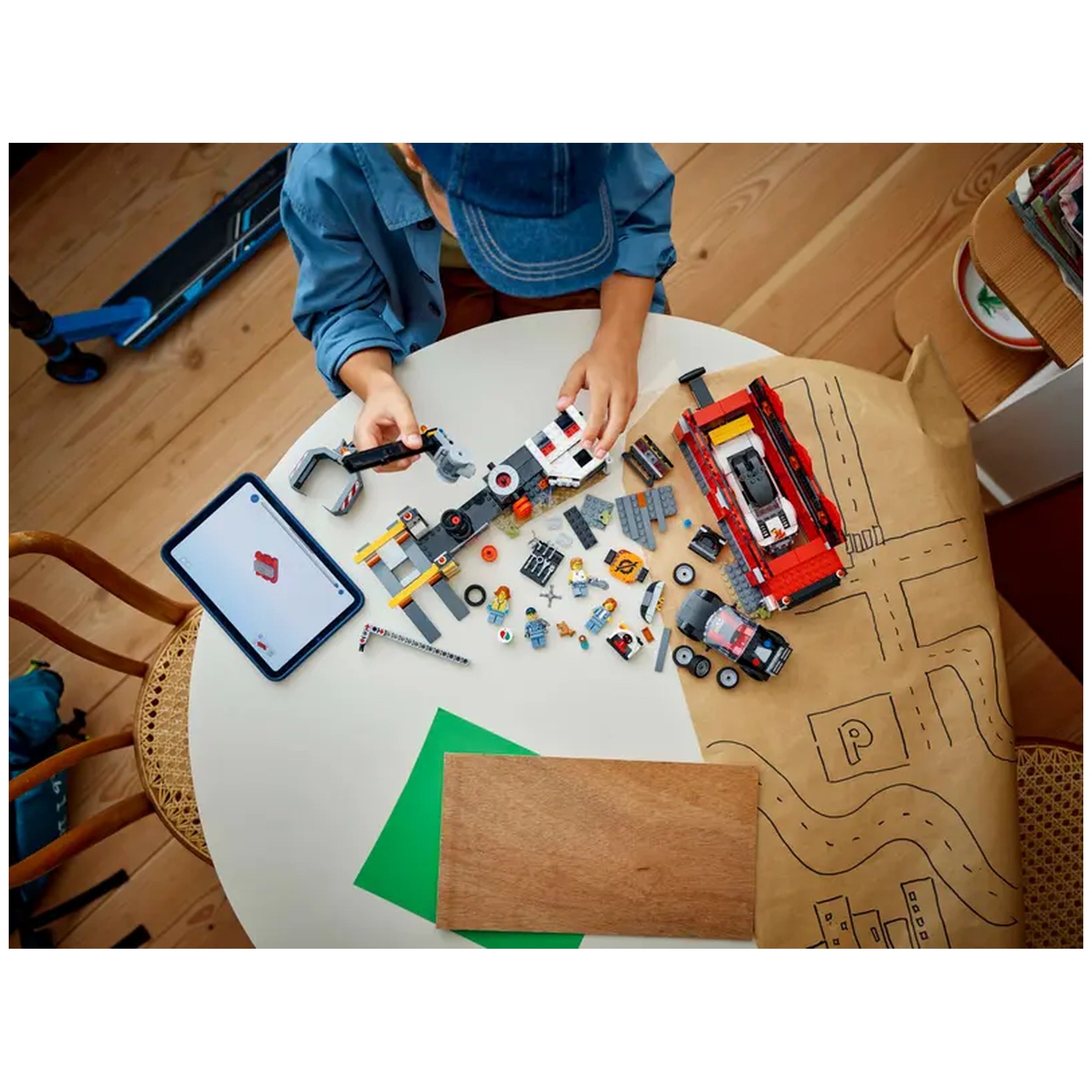 Child assembling a LEGO City Scrapyard set on a table, surrounded by colorful bricks, tools, two toy cars, and a digital tablet displaying instructions.