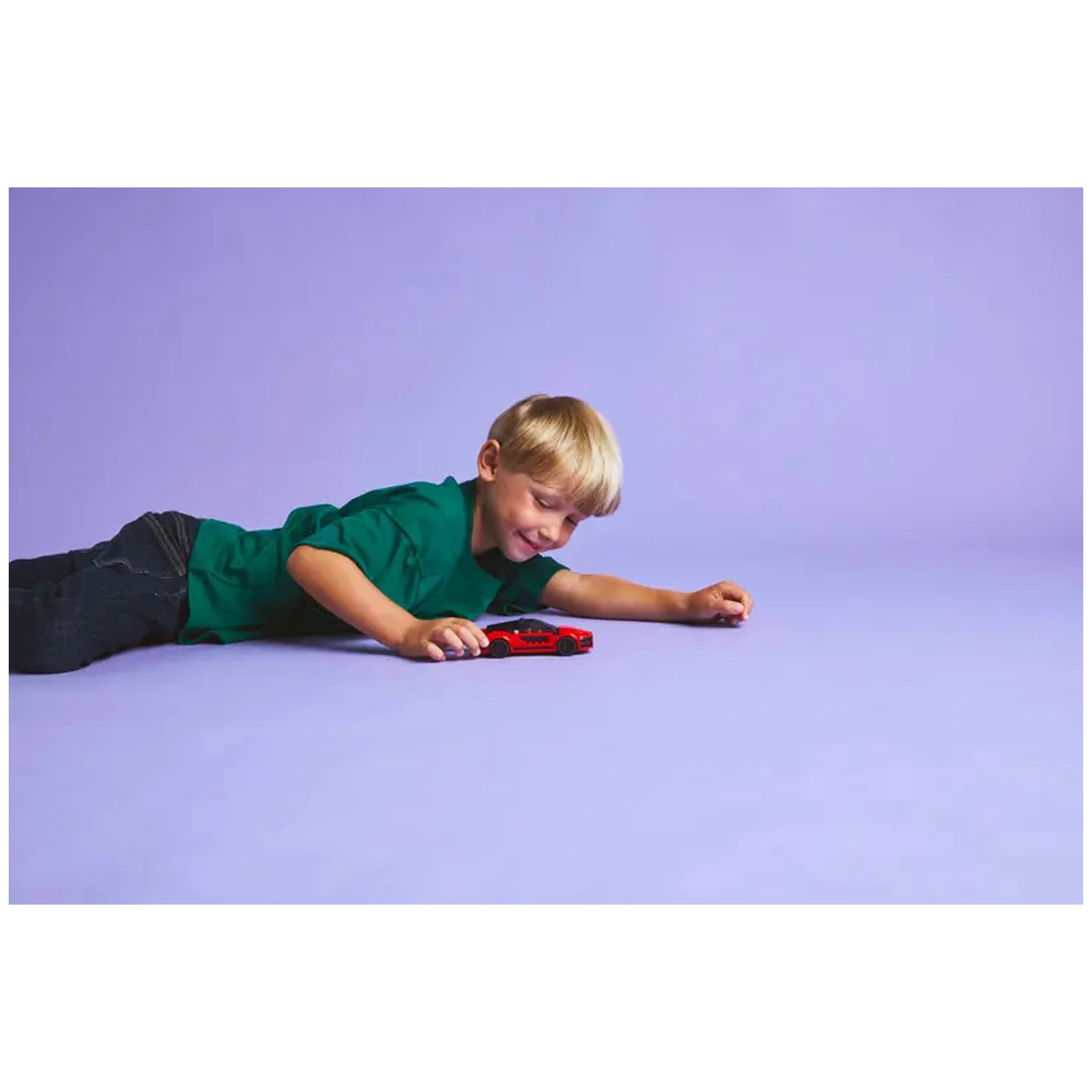 A young boy lies on a purple floor, smiling as he plays with a bright red LEGO City sports car. He reaches towards the toy, embodying excitement and imagination.
