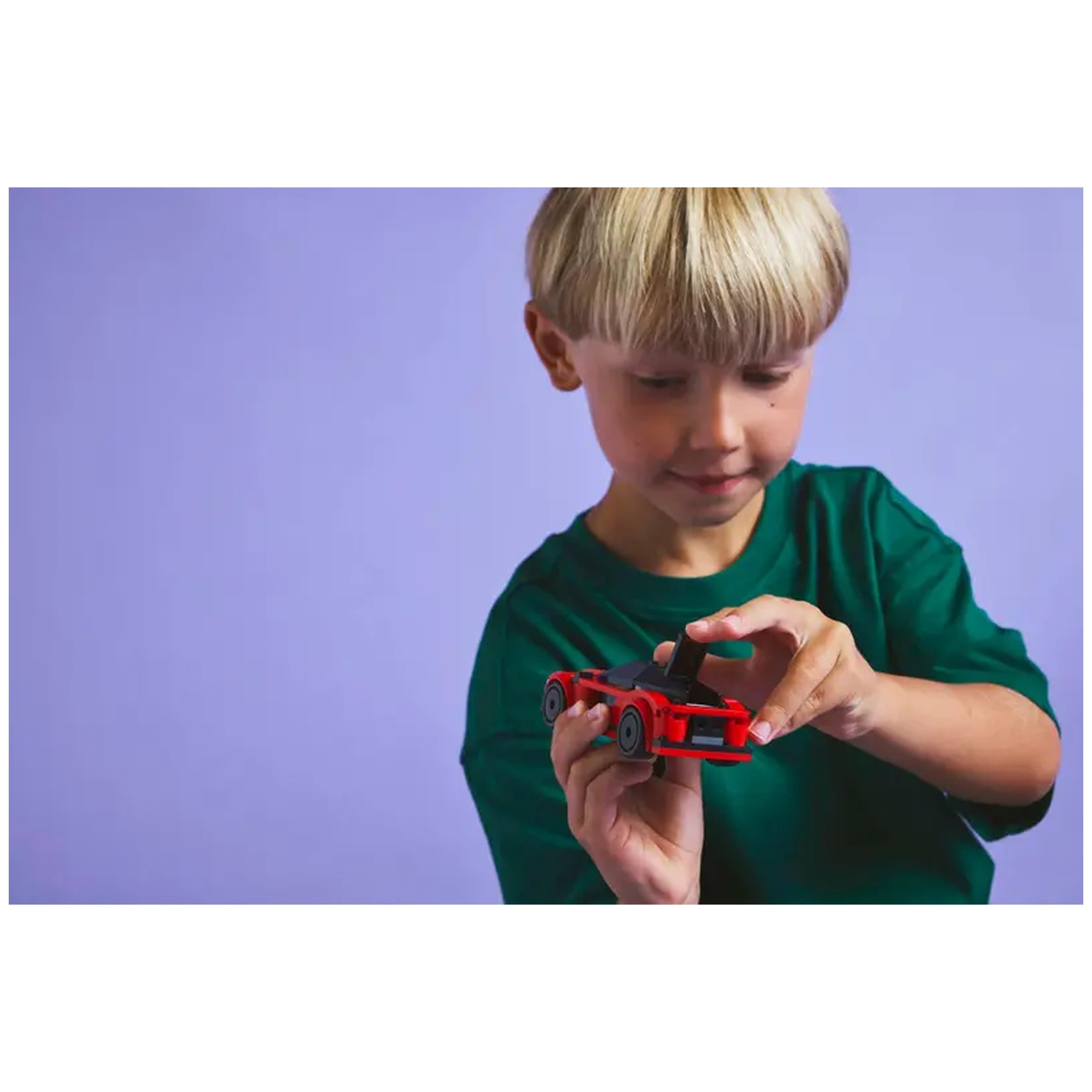 A young child in a green shirt carefully examines a bright red LEGO® sports car against a soft purple background.