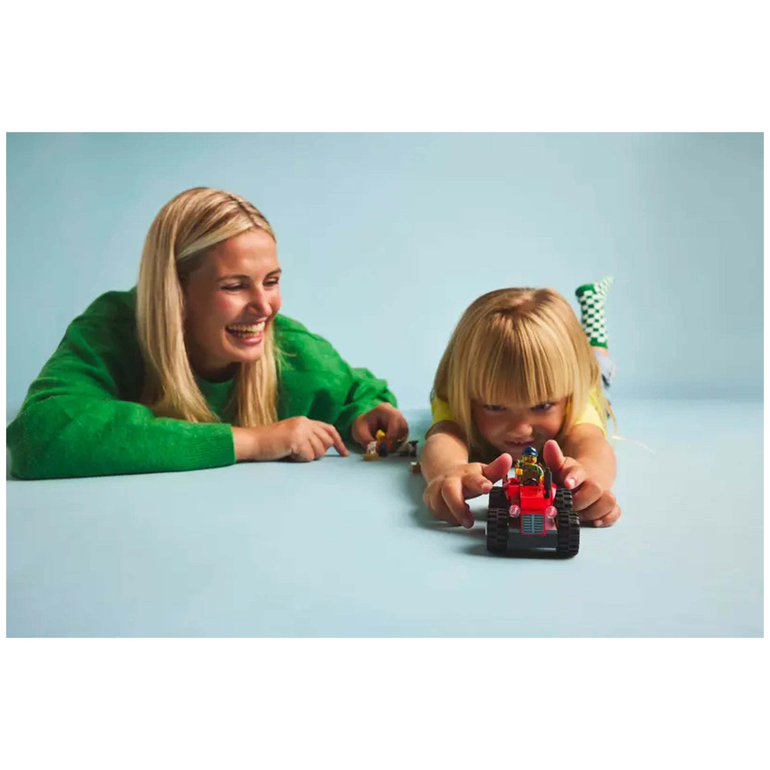 Smiling adult and child play on a blue surface, focused on a vibrant red LEGO City tractor set, engaging in imaginative play.