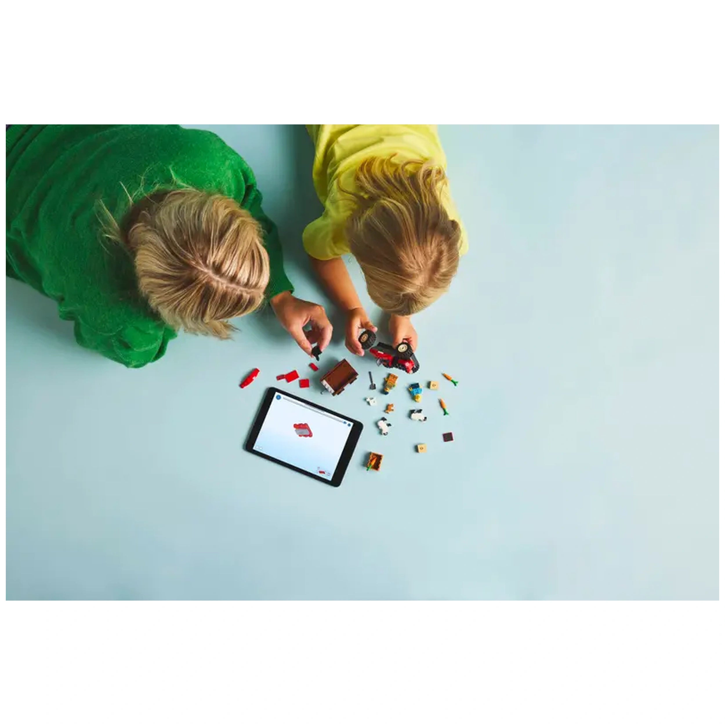 A child in a yellow shirt and an adult in a green sweater play with LEGO pieces, including a red tractor, on a light blue surface beside a tablet.