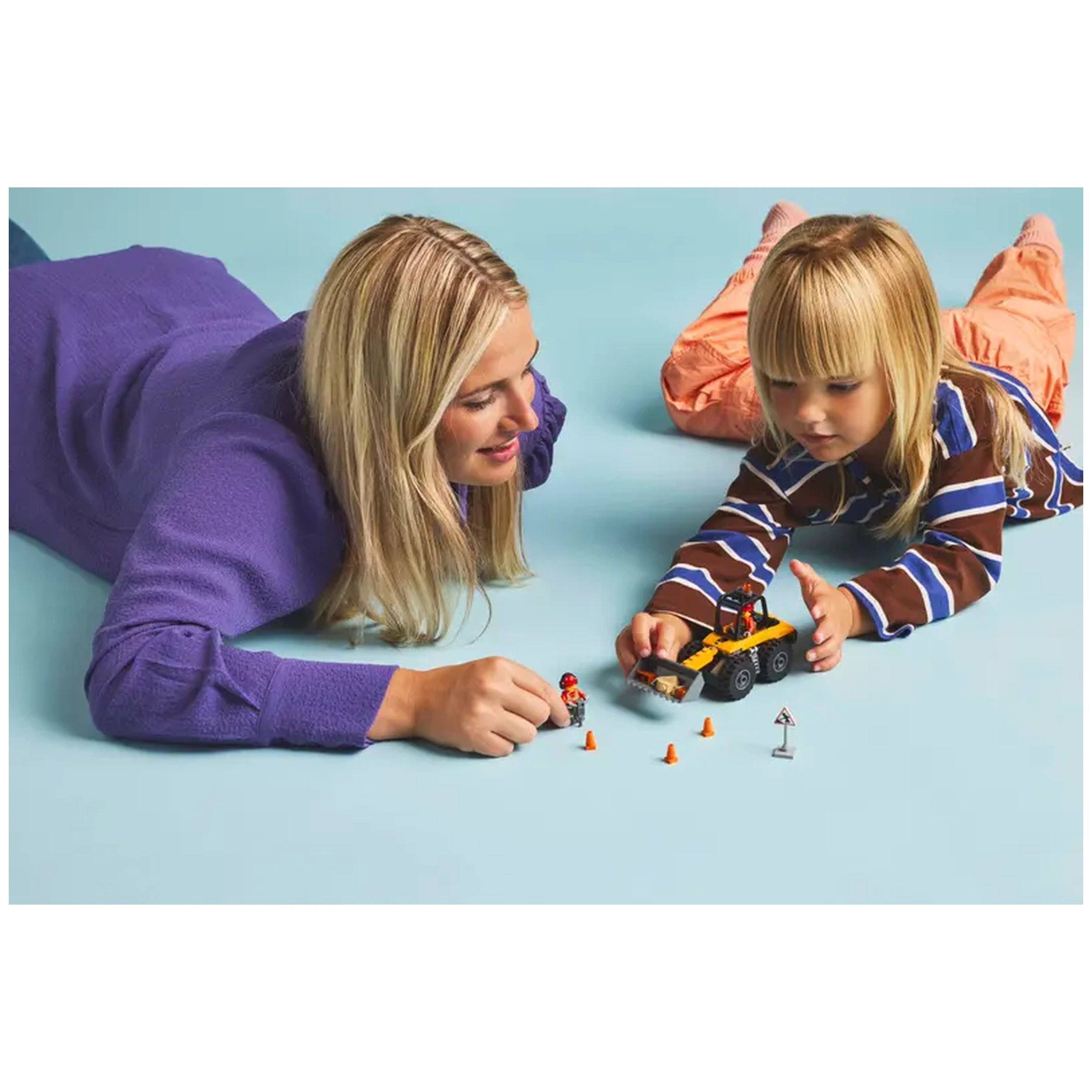 A woman and a child play together on a light blue floor, engaging with a LEGO® City Wheel Loader set. The child, wearing a striped shirt, moves the yellow construction vehicle, while the woman, in a purple sweater, assists. Several small construction accessories, including cones and roadwork signs, are arranged around them.