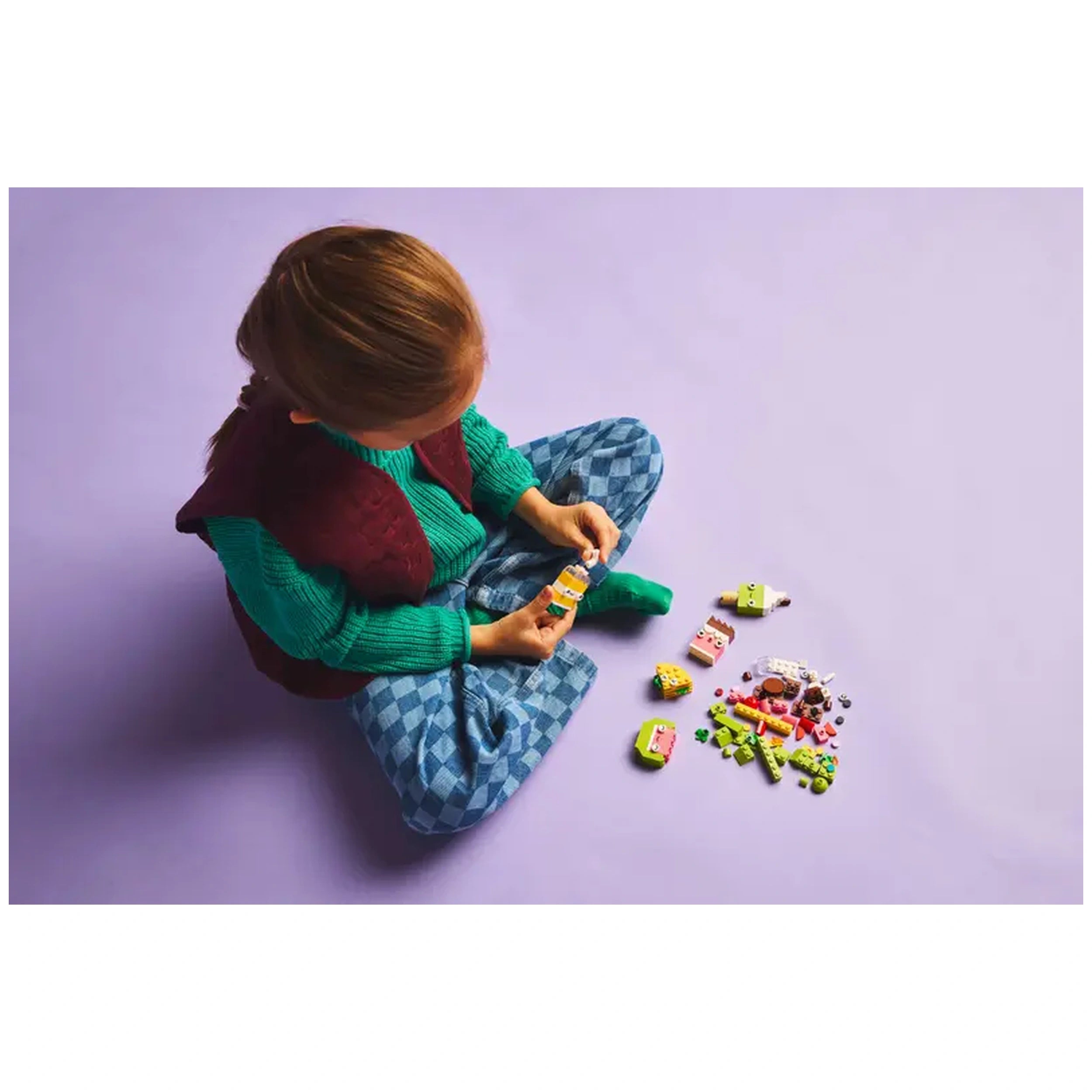 A child sits cross-legged on a purple surface, focused on assembling colorful LEGO bricks into food characters. Surrounding them are various vibrant pieces, including cupcakes and tacos, showcasing imaginative play. The child wears a teal sweater and maroon vest, her brown hair cascading lightly.