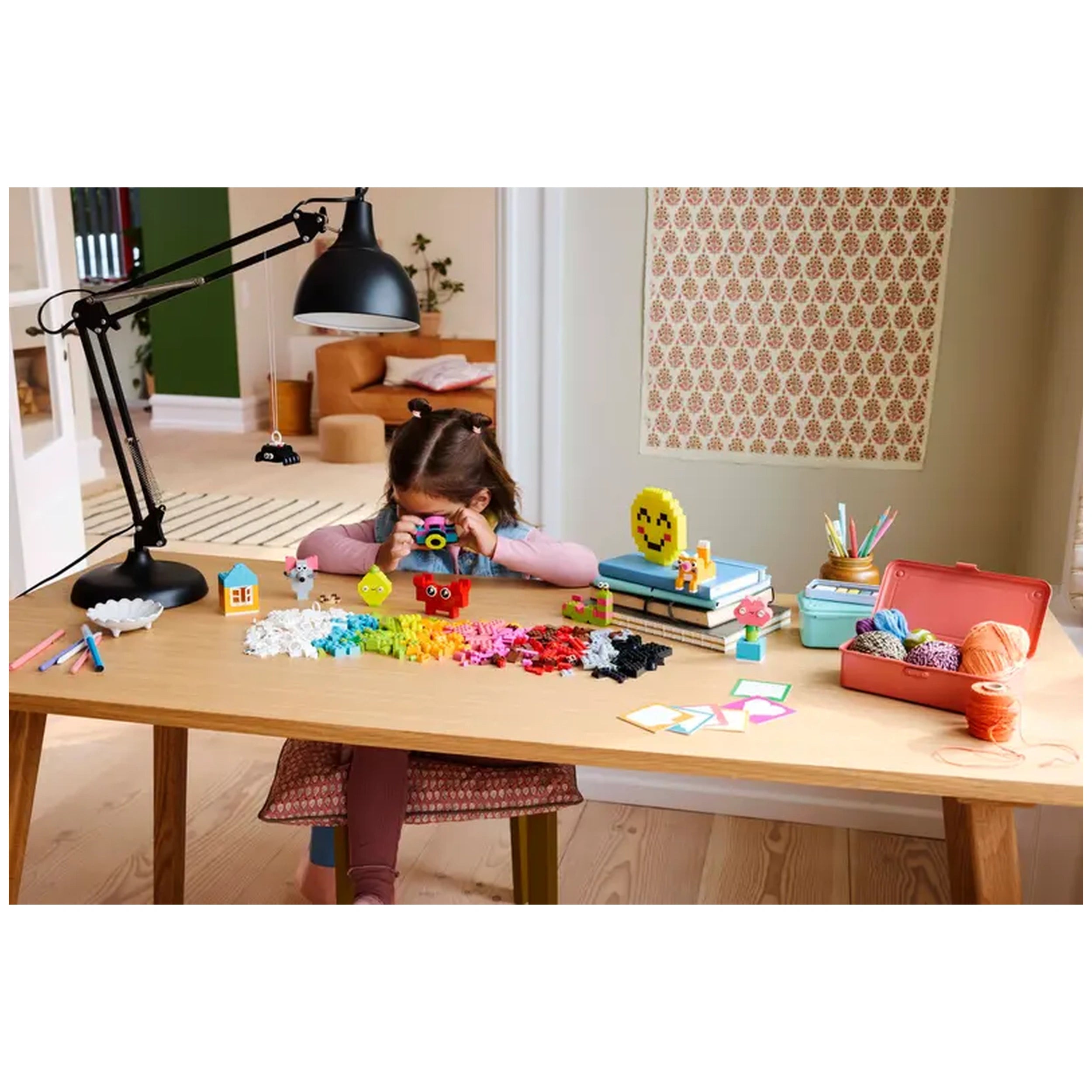 A young girl sits at a wooden table, focusing on a colorful LEGO model while surrounded by scattered bricks in vibrant hues. A black desk lamp illuminates her creative space, which includes craft supplies, a smiley face decoration, and a pink box filled with yarn. An inviting room with a patterned wall can be seen in the background.