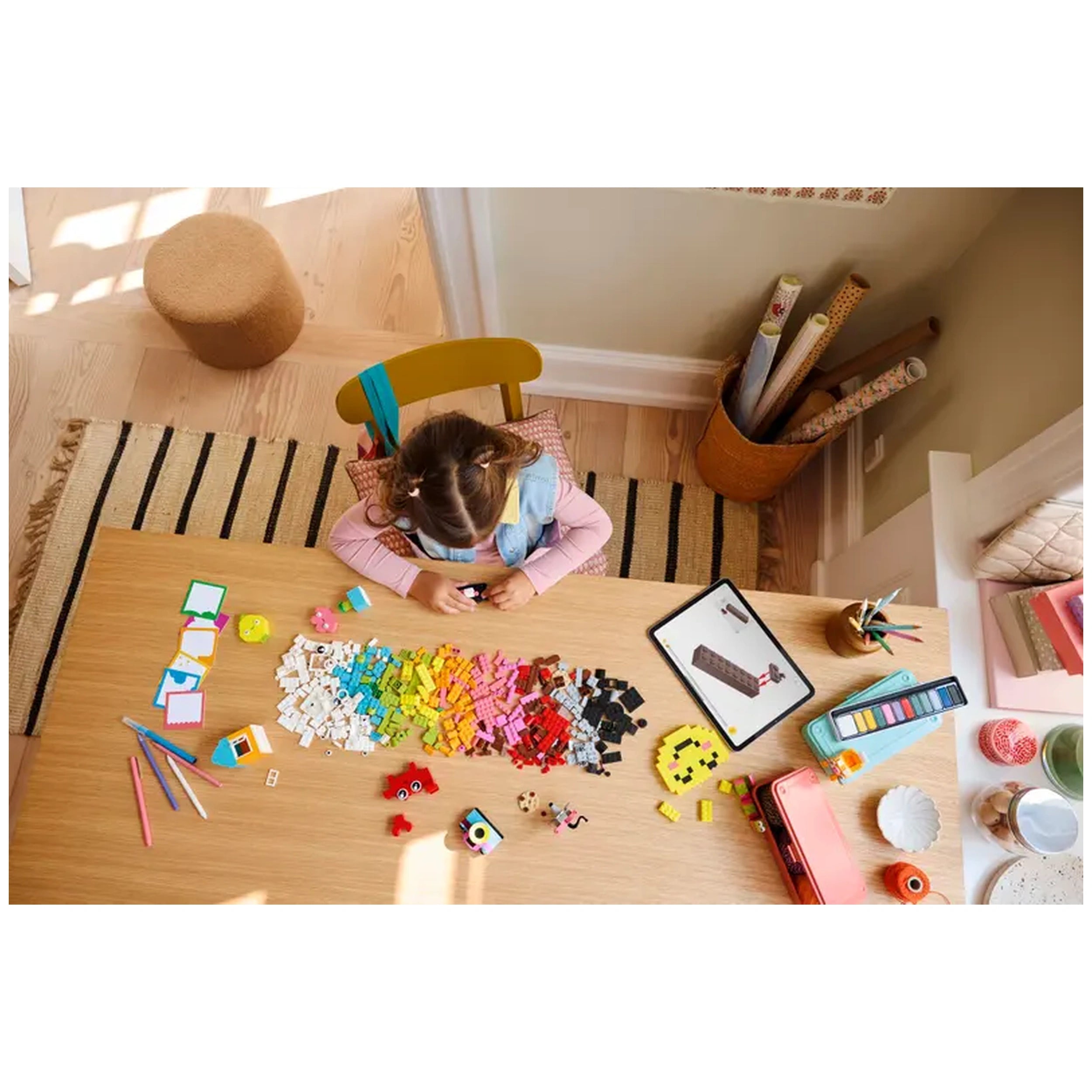 A young girl sits at a wooden table covered with colorful LEGO bricks and play items. She concentrates on building while surrounded by vibrant pieces, a tablet displaying a model nearby, and various art supplies. A patterned rug and a cozy stool add warmth to the creative space.