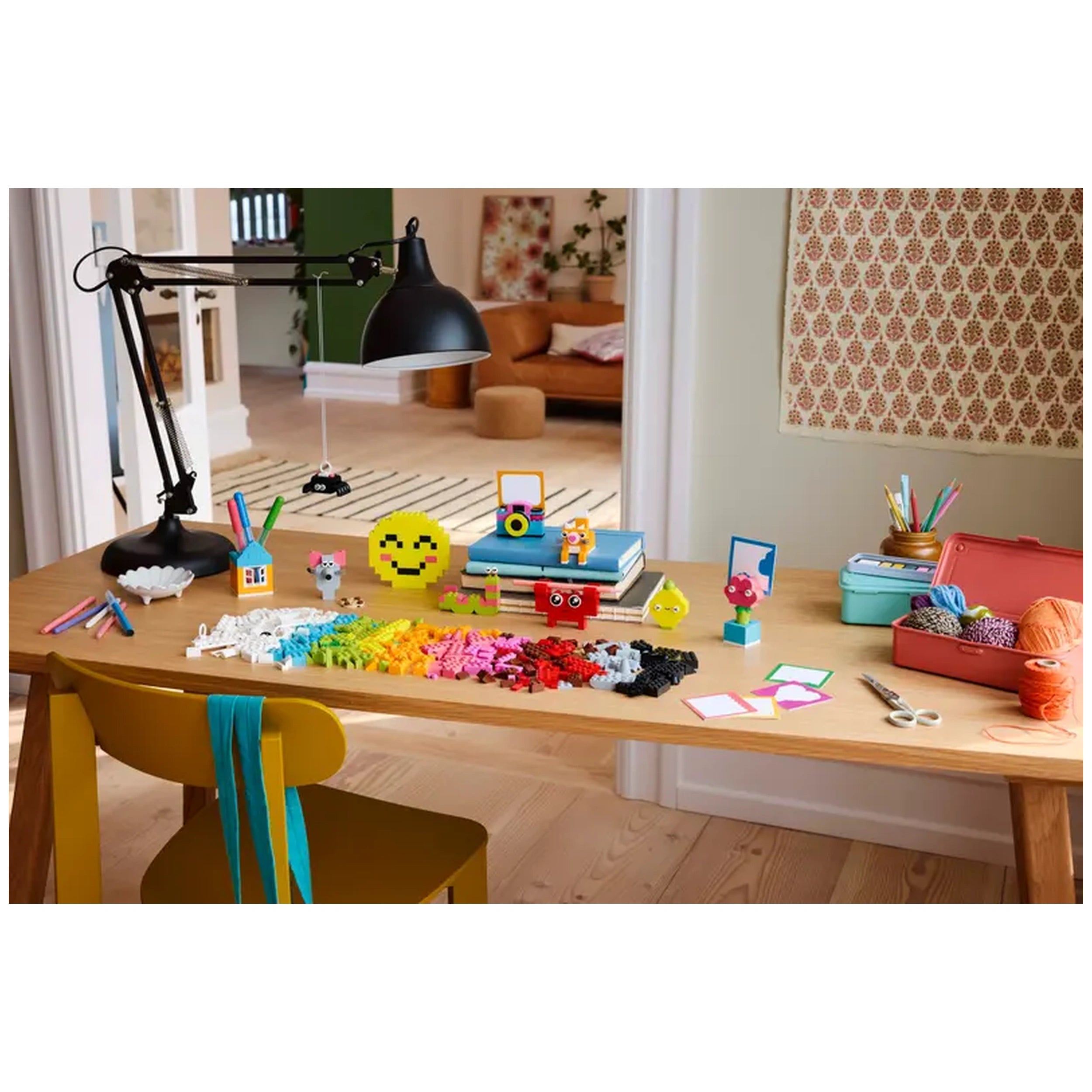 A brightly lit wooden table filled with colorful LEGO bricks, markers, and playful creations. A smiling emoji figure sits among red and yellow toys, next to vibrant building elements. A black desk lamp illuminates the scene, with a stack of books in the background and a soft couch visible. Scissors and craft supplies are scattered, showcasing a fun and creative workspace for kids aged 5 and up.