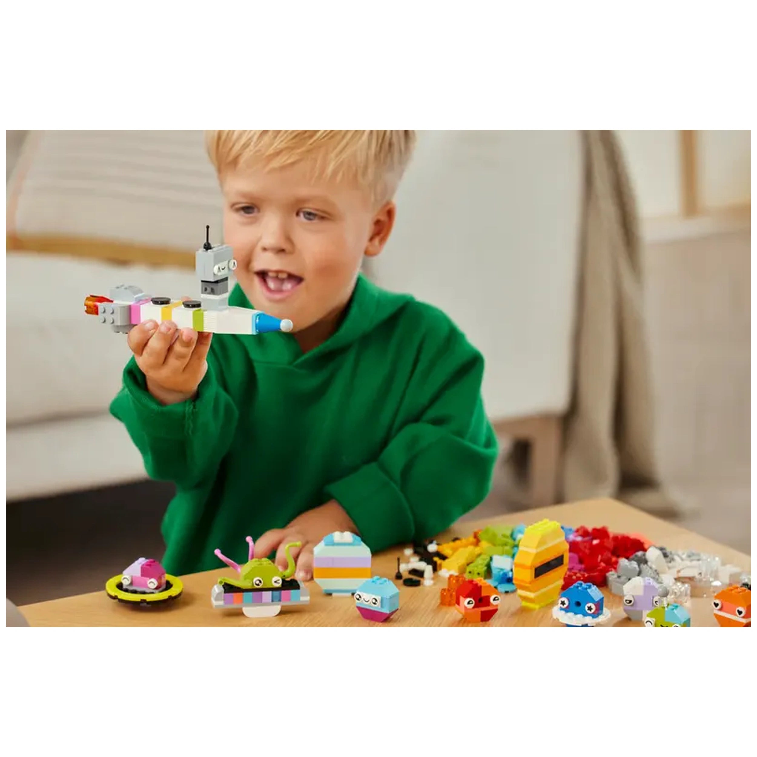 A young boy in a green hoodie joyfully holds a colorful LEGO spacecraft. Surrounding him are various space-themed LEGO creations, including an alien, planets, and vibrant bricks on a table.