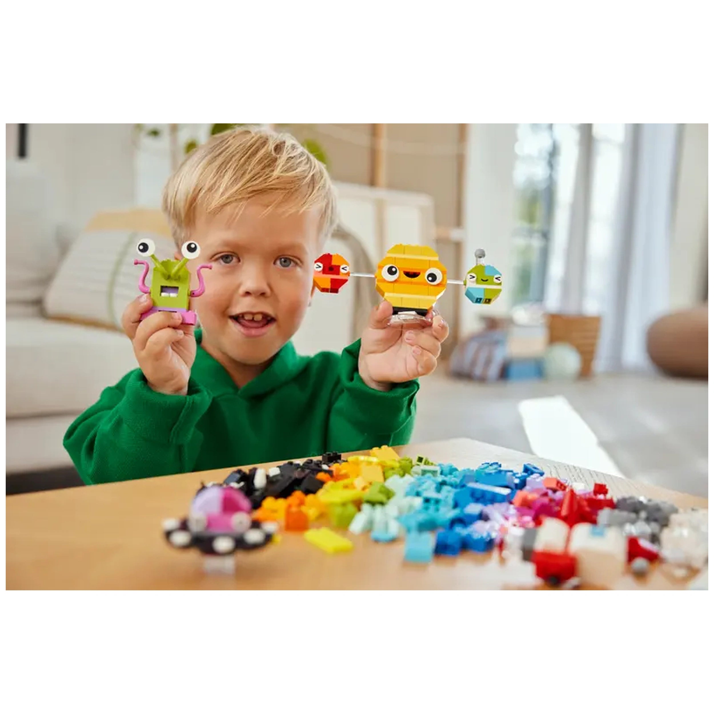 Young boy wearing a green hoodie smiles while holding colorful LEGO characters, surrounded by a vibrant assortment of LEGO bricks.