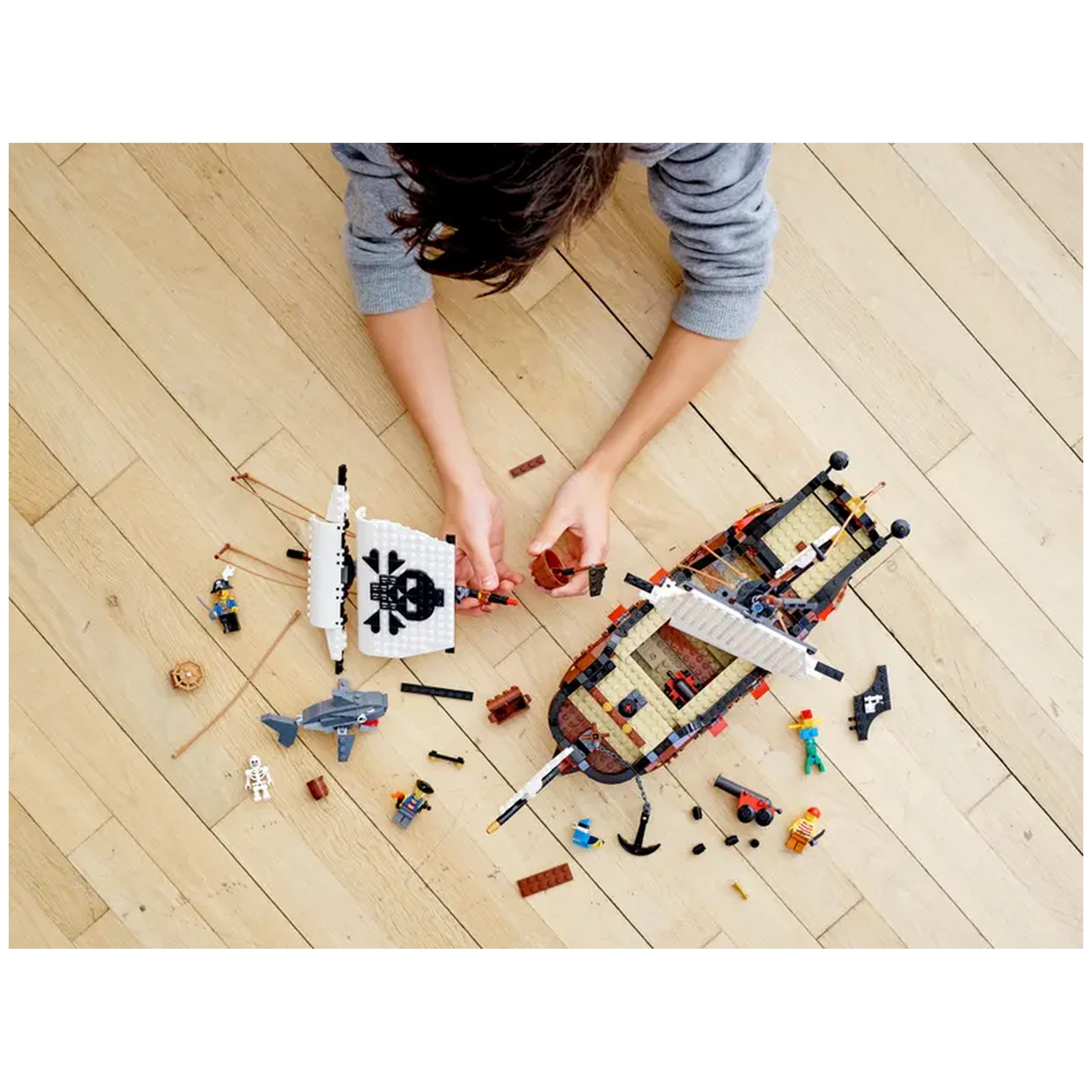 Child assembling the LEGO® Creator 3in1 Pirate Ship on a wooden floor, surrounded by minifigures, accessories, and detailed ship parts.