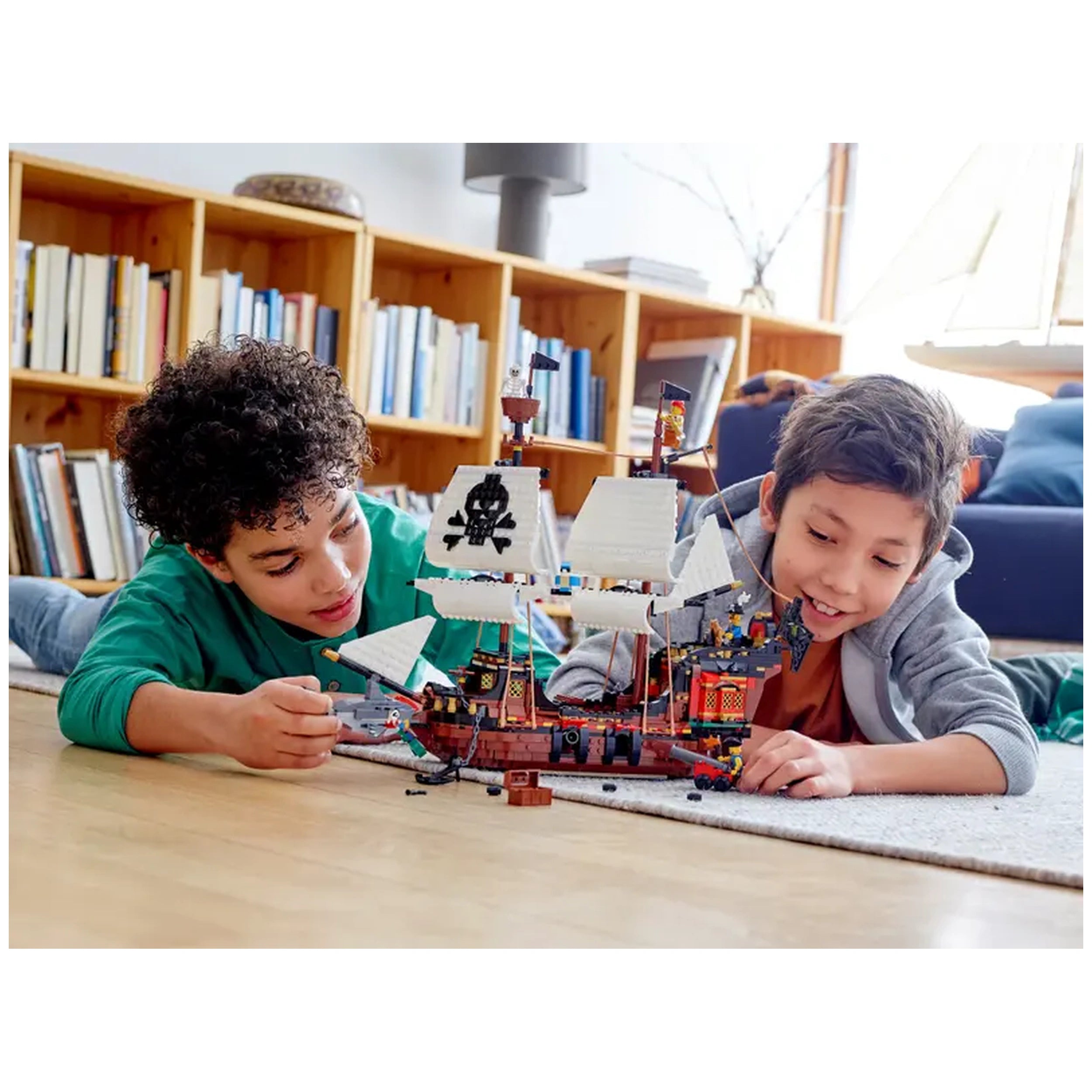 Two children play together on the floor, focusing intently on a detailed LEGO® Creator 3in1 Pirate Ship. The ship features white sails, a skull and crossbones flag, and colorful deck elements. Bookshelves filled with books are visible in the background, creating a cozy indoor environment.