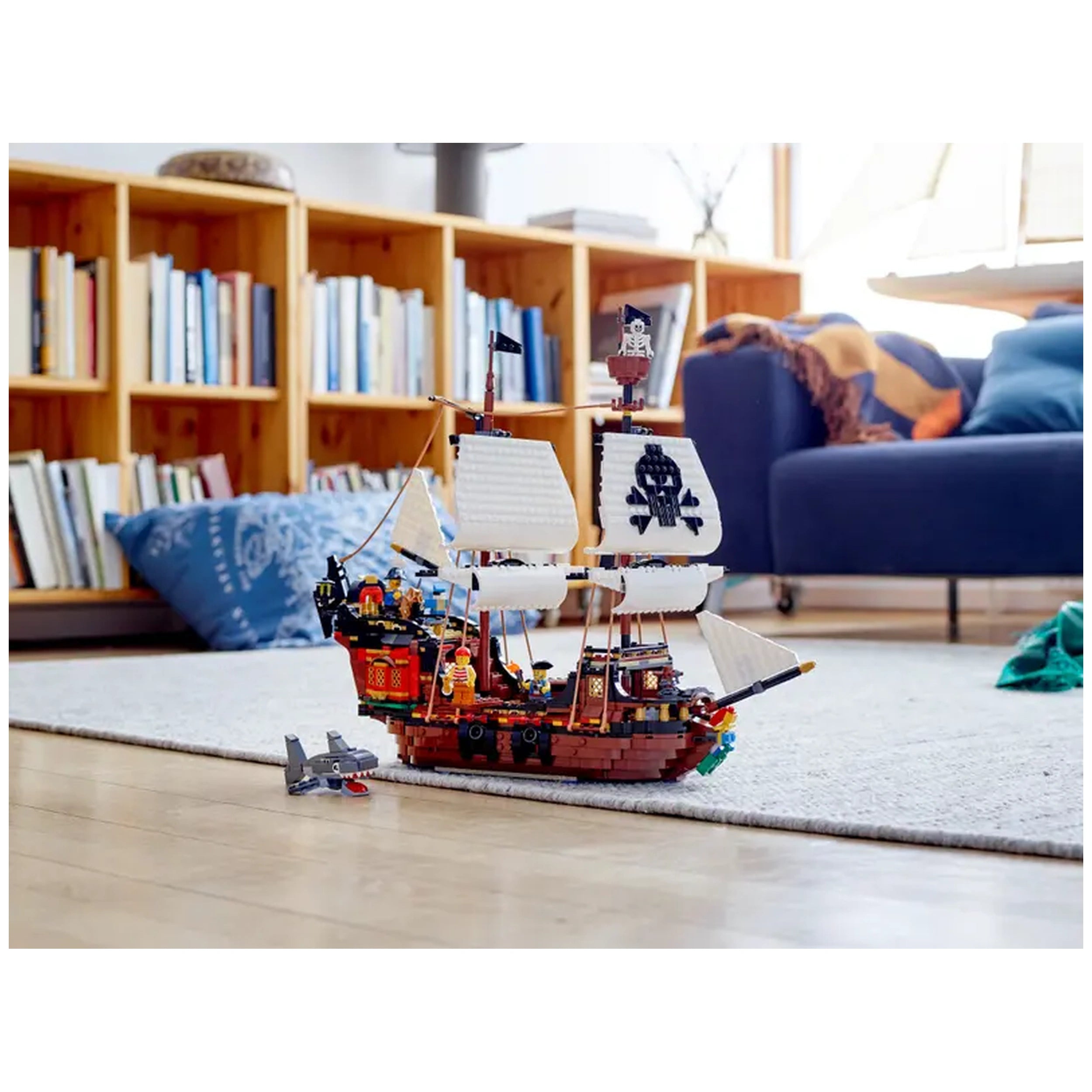 Colorful LEGO® Creator 3in1 Pirate Ship on a light wooden floor, surrounded by a cozy living room with bookshelves and a blue couch. A shark figure lies nearby.
