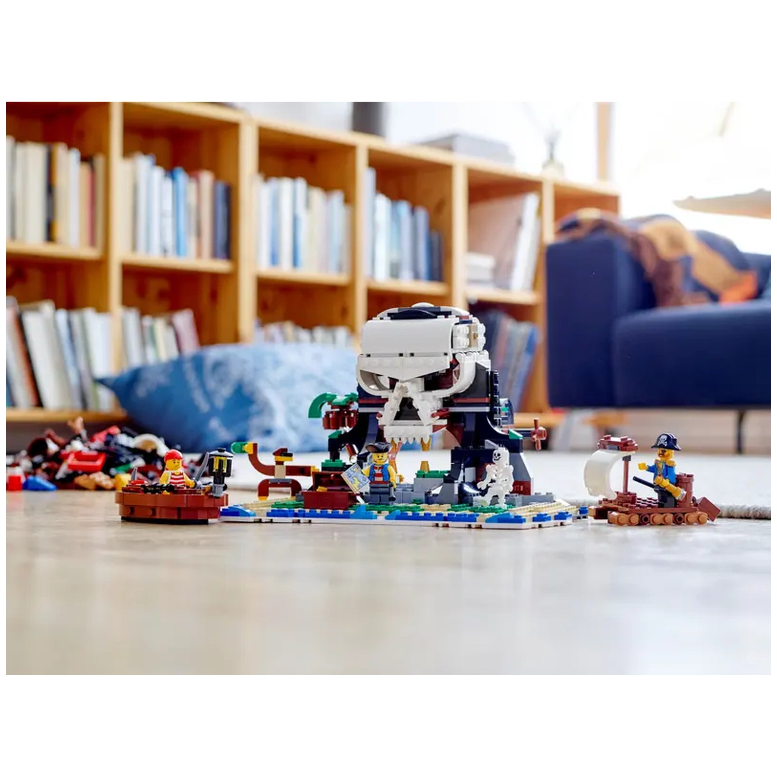 A colorful LEGO® Creator 3in1 Pirate Ship set featuring a pirate ship with sails, cannons, and three minifigures. The scene includes a skull-shaped island and buildable figures like a shark, surrounded by a wooden boat. In the background, a cozy room with bookshelves and a couch is visible.