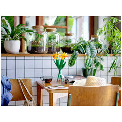 LEGO® Creator Daffodils construction set displayed on a wooden table, featuring two yellow and two white flowers in a vase, surrounded by green plants and a cup of tea.