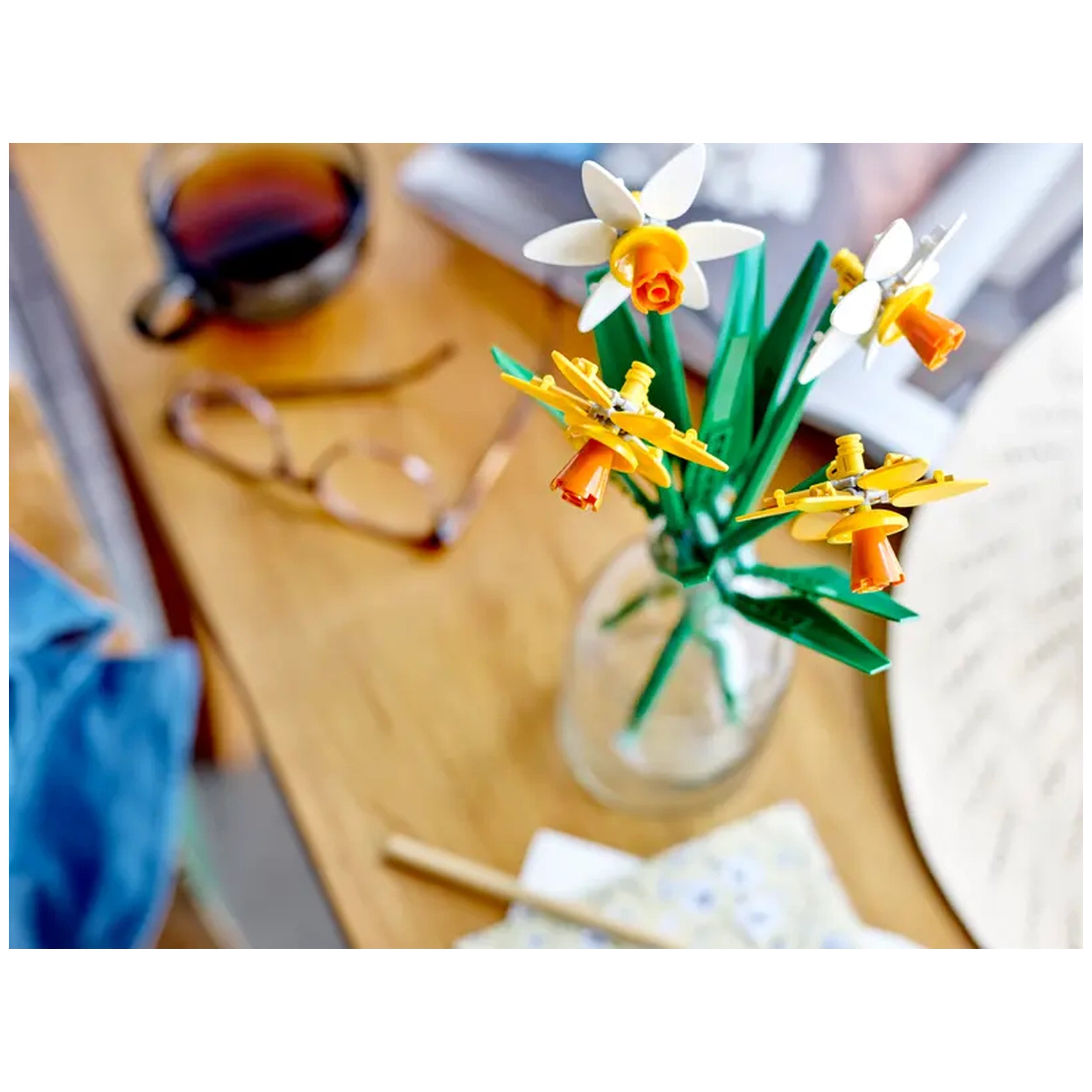 A colorful arrangement of LEGO® Daffodils in a glass vase, featuring two yellow flowers and two white flowers with green stems, set on a wooden table beside a cup of coffee and reading glasses.