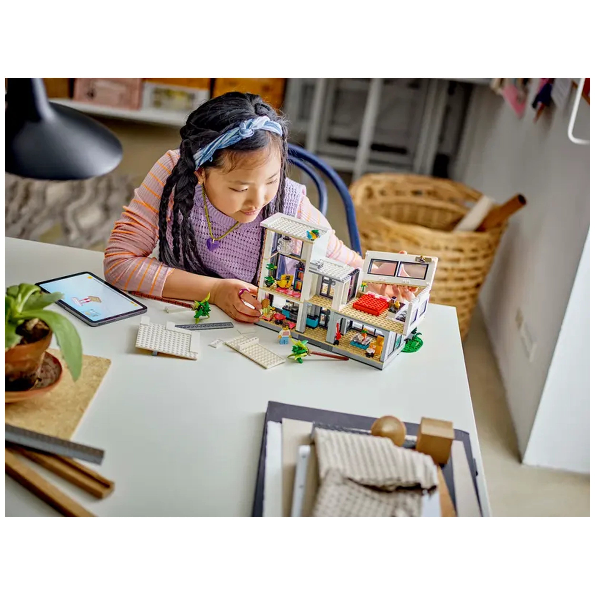 A young girl with braided hair builds a colorful LEGO Creator Modern House set on a bright table, surrounded by LEGO pieces and plant, focusing intently on her construction.