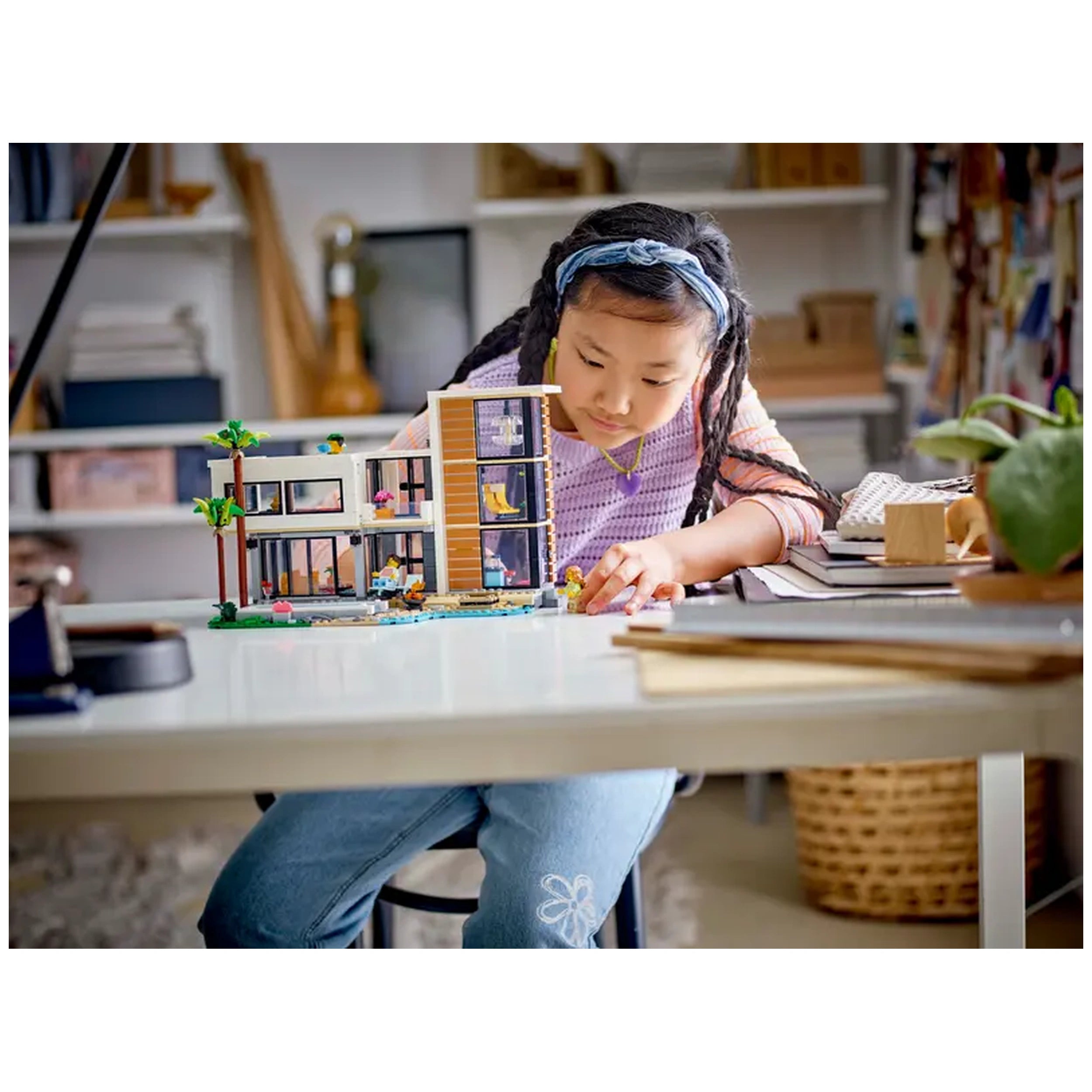 A young girl with braided hair focuses intently on the LEGO Creator Modern House construction set, exploring its colorful features on a white table. The stylish model showcases a two-story design with modern furnishings and palm trees, enhancing imaginative play. Books and a plant are visible in the background, creating a cozy atmosphere.