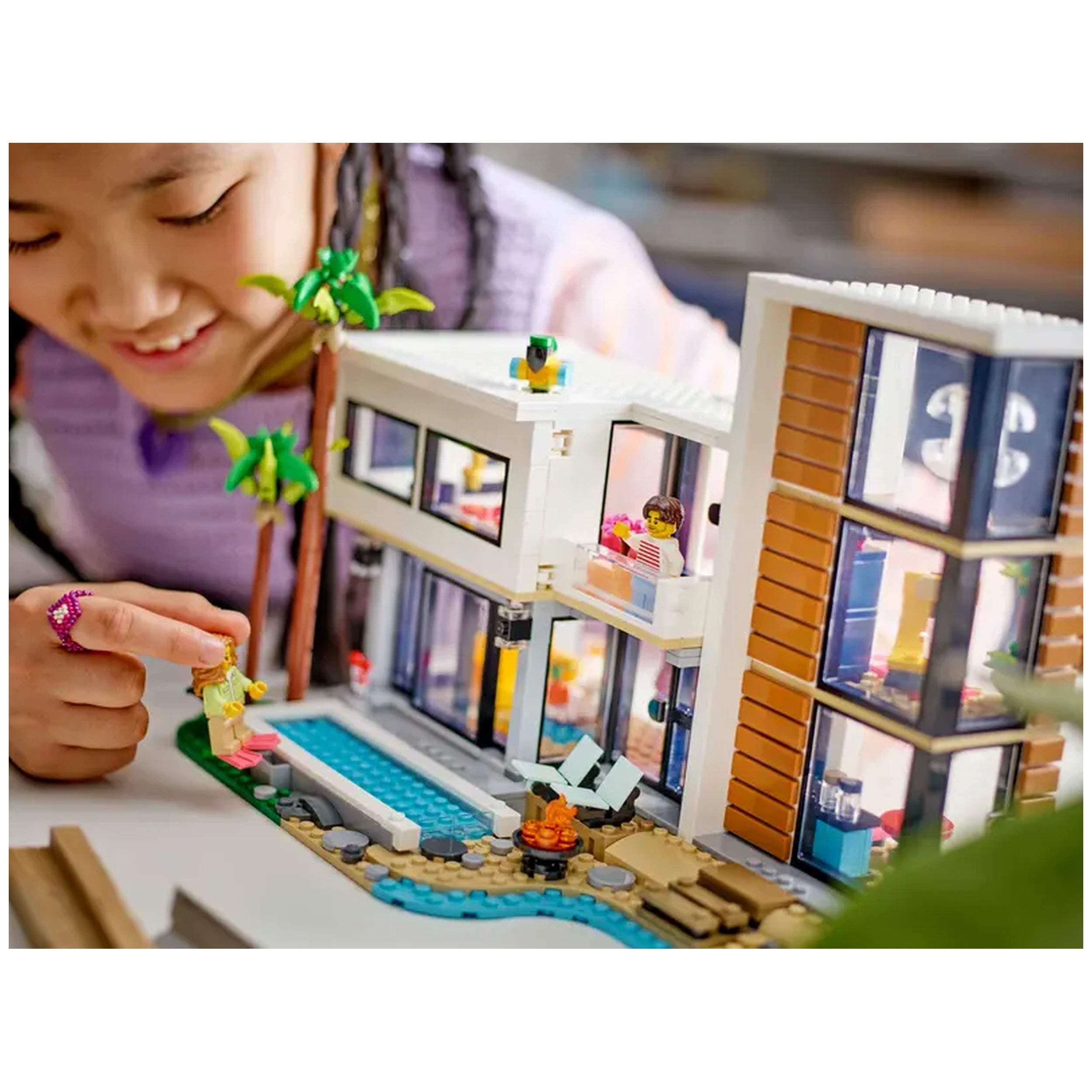 A child leans over a stylish LEGO Creator Modern House set, pointing excitedly at a minifigure by a swimming pool surrounded by palm trees.