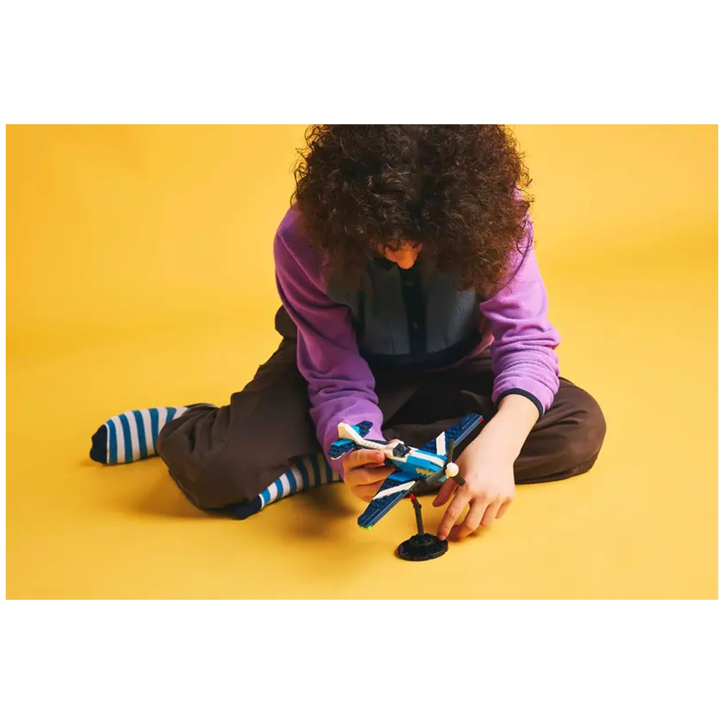 A child sits on a yellow background, focused on assembling a blue and white LEGO race plane, with vibrant striped socks visible.