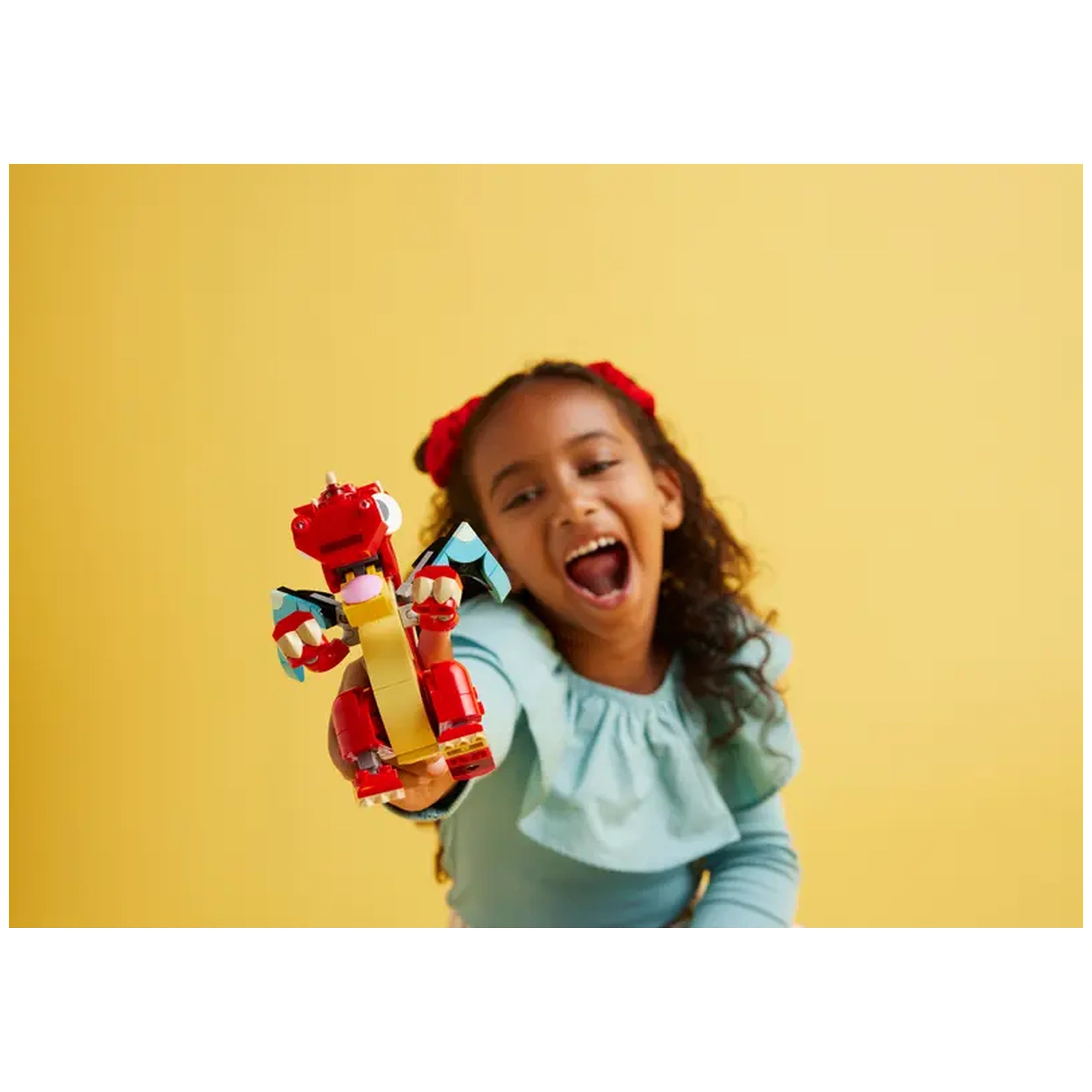 A smiling girl with curly hair holds up a vibrant red LEGO Creator dragon against a sunny yellow background, showcasing her excitement.