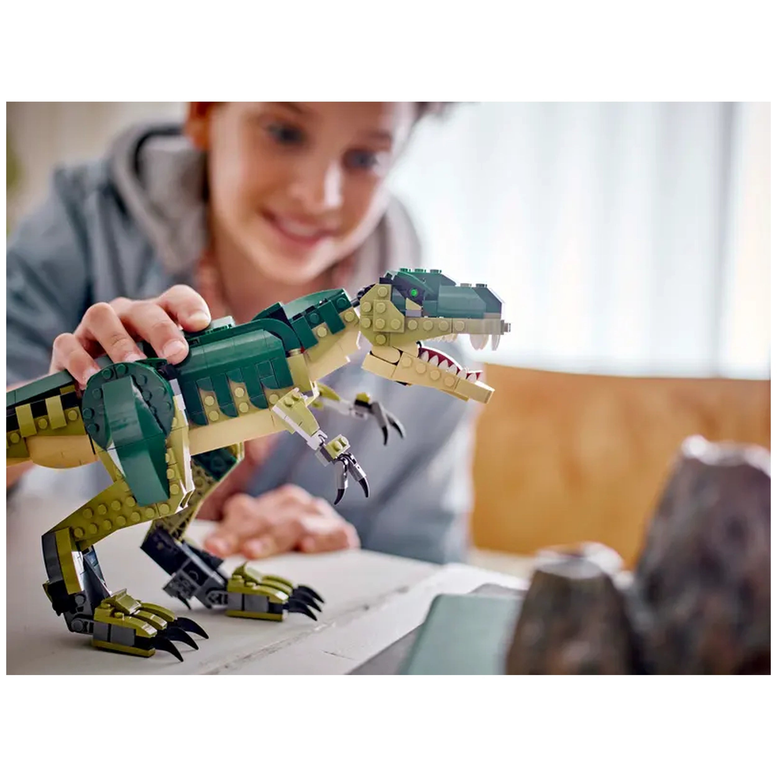 A child smiles while playfully manipulating a green and yellow LEGO T. rex, showcasing its articulated limbs and fierce expression, with a blurred background of play elements.