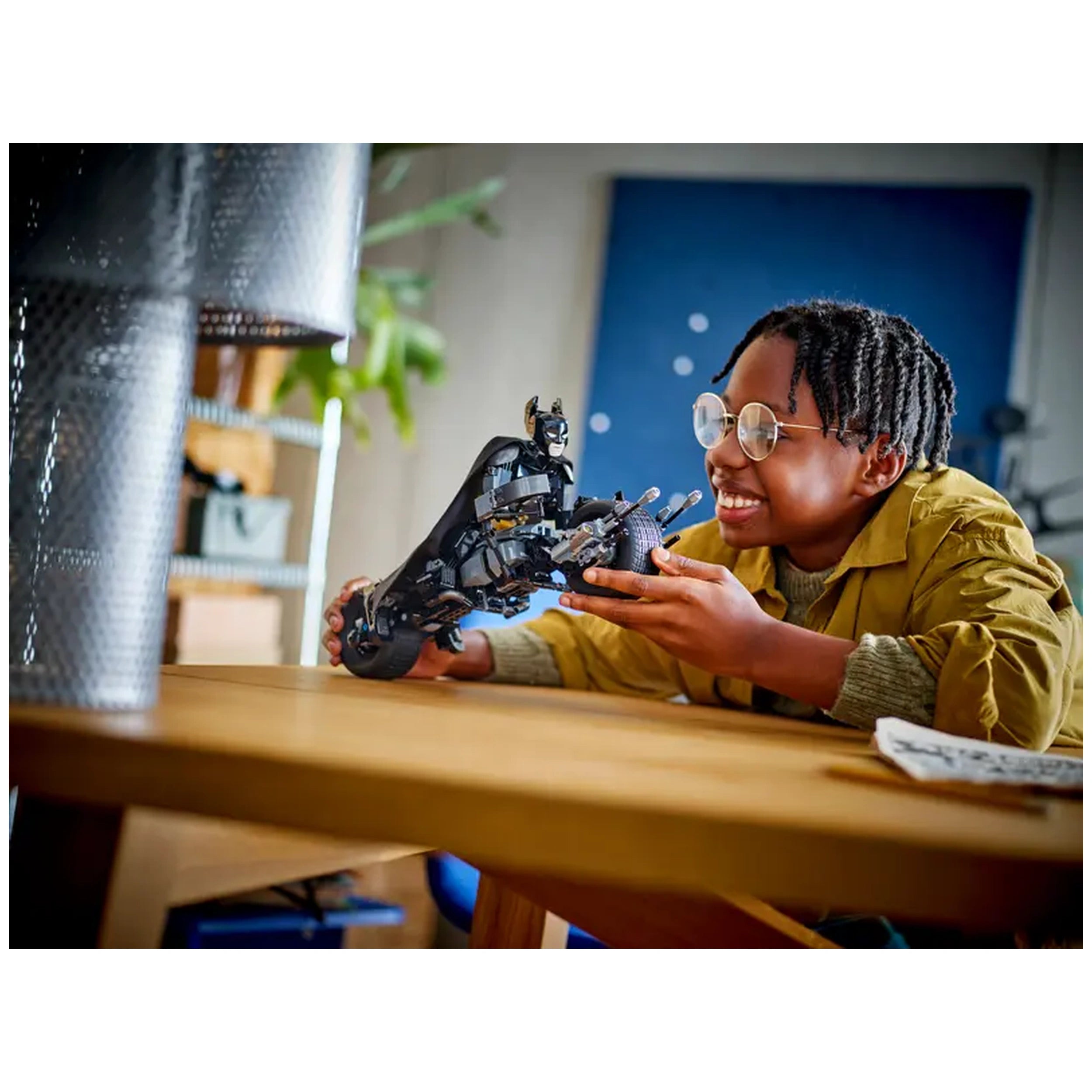 A child wearing glasses smiles while holding a LEGO Batman construction figure and Bat-Pod bike, seated at a wooden table.