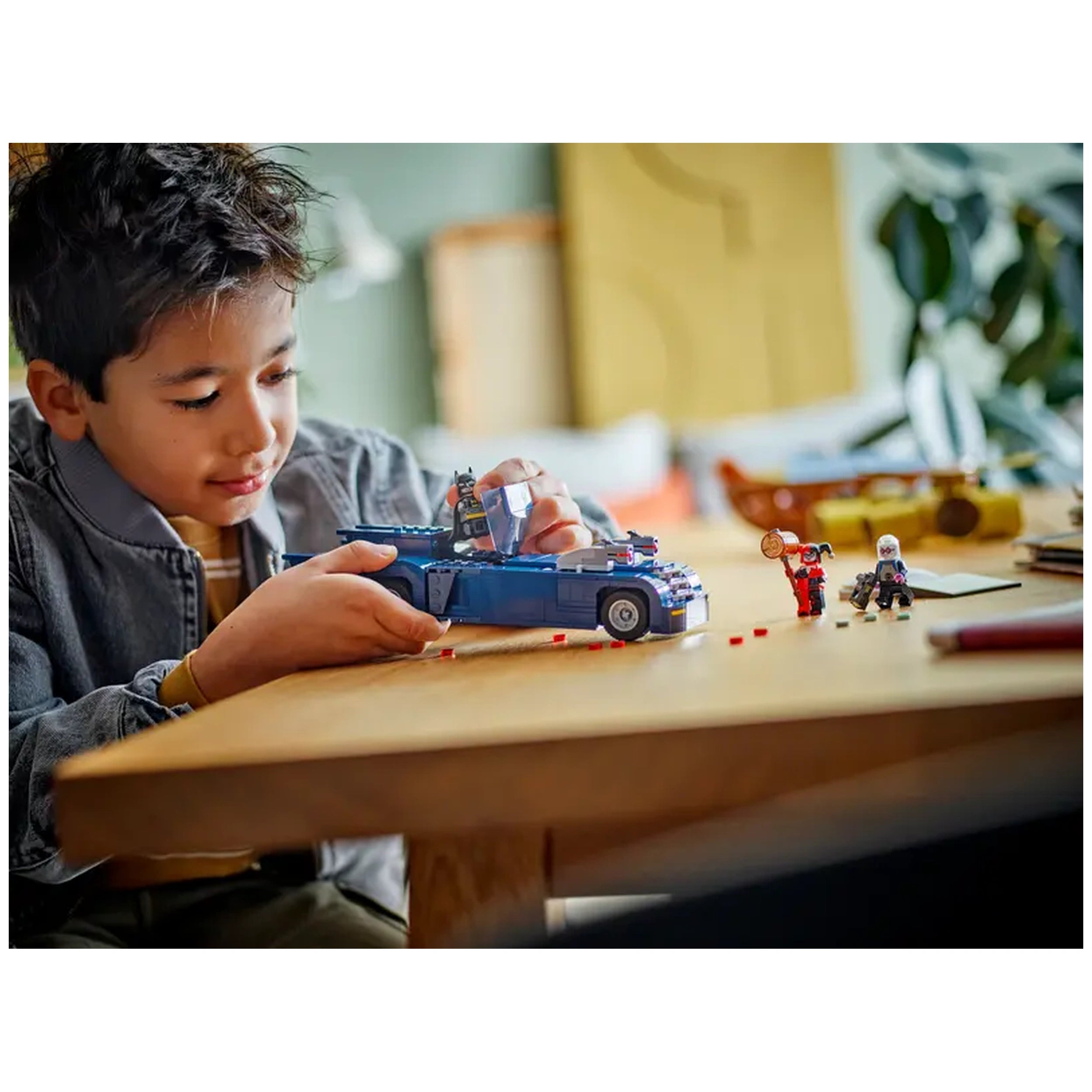 A young boy carefully assembles the blue Batmobile from the LEGO DC Comics set, holding a Batman minifigure while two other characters, Harley Quinn and Mr. Freeze, stand nearby on a wooden table.