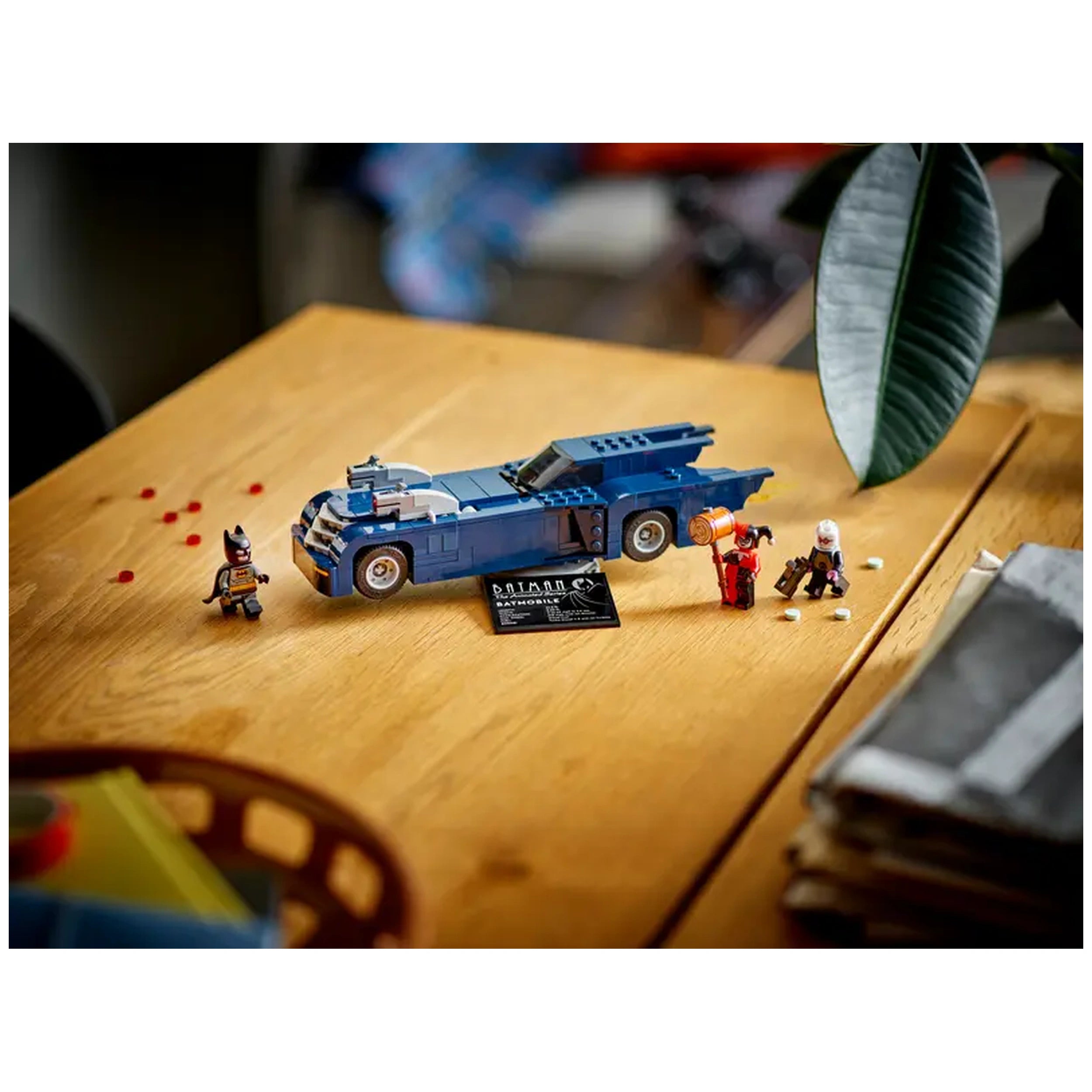Batman minifigure stands next to the blue Batmobile, with Harley Quinn and Mr. Freeze minifigures on a wooden table, featuring a Batman logo card.