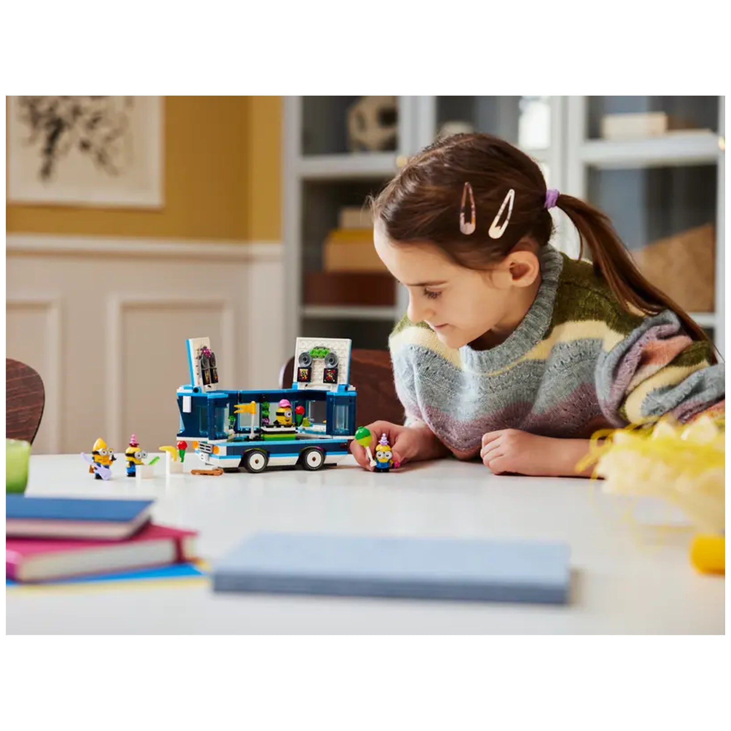 Young girl in a colorful sweater plays with the LEGO® Minions' Music Party Bus set on a table, surrounded by Minion figures and building blocks.