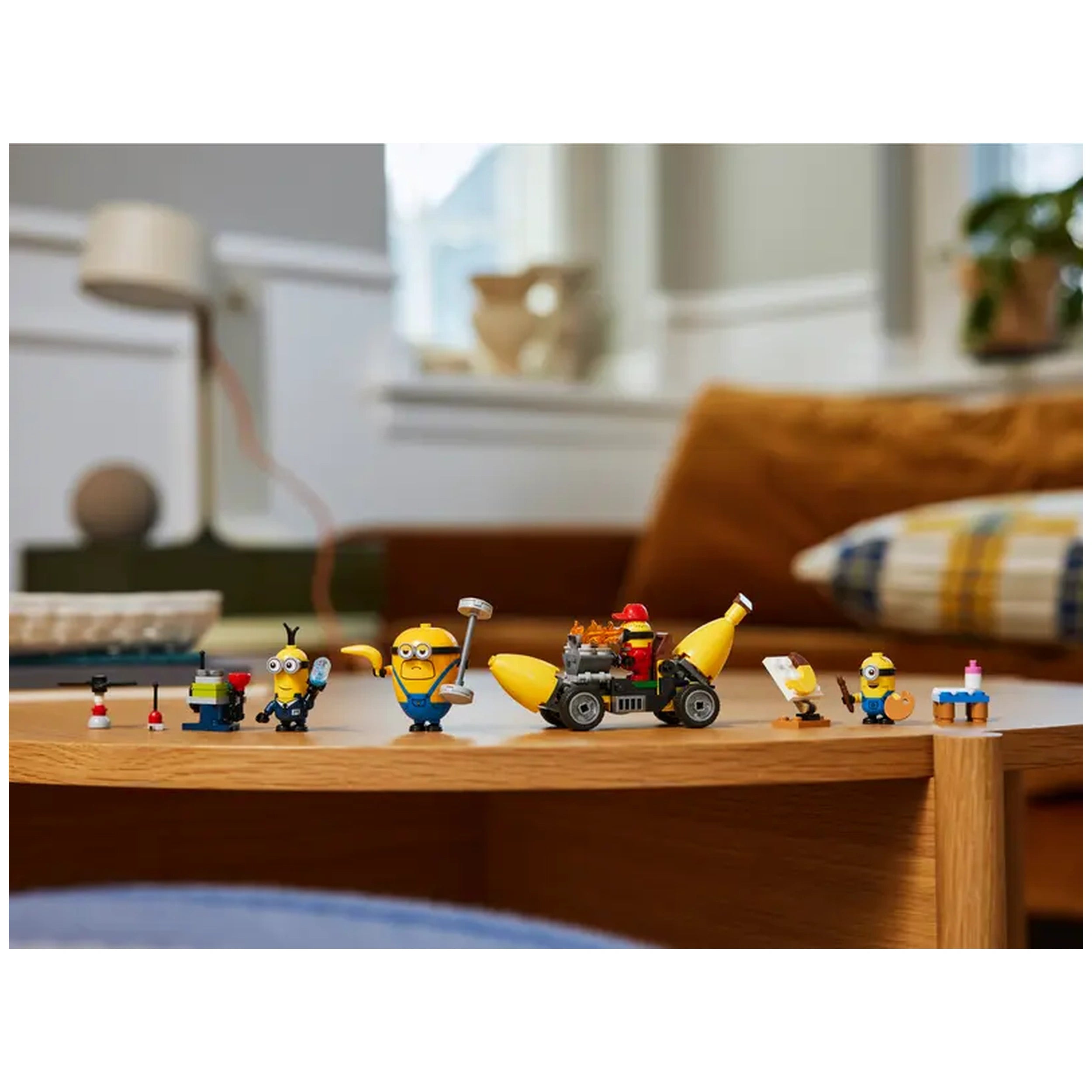 Four LEGO® Minion figures and a vibrant yellow banana car sit on a wooden table. The figures include Mega Minion Dave, Mel with a paintbrush, Agent AVL Minion Tim, and Pit Crew Minion Ron, each showcasing distinct accessories. The banana car features a removable engine and vibrant details, set against a cozy background with soft furnishings.