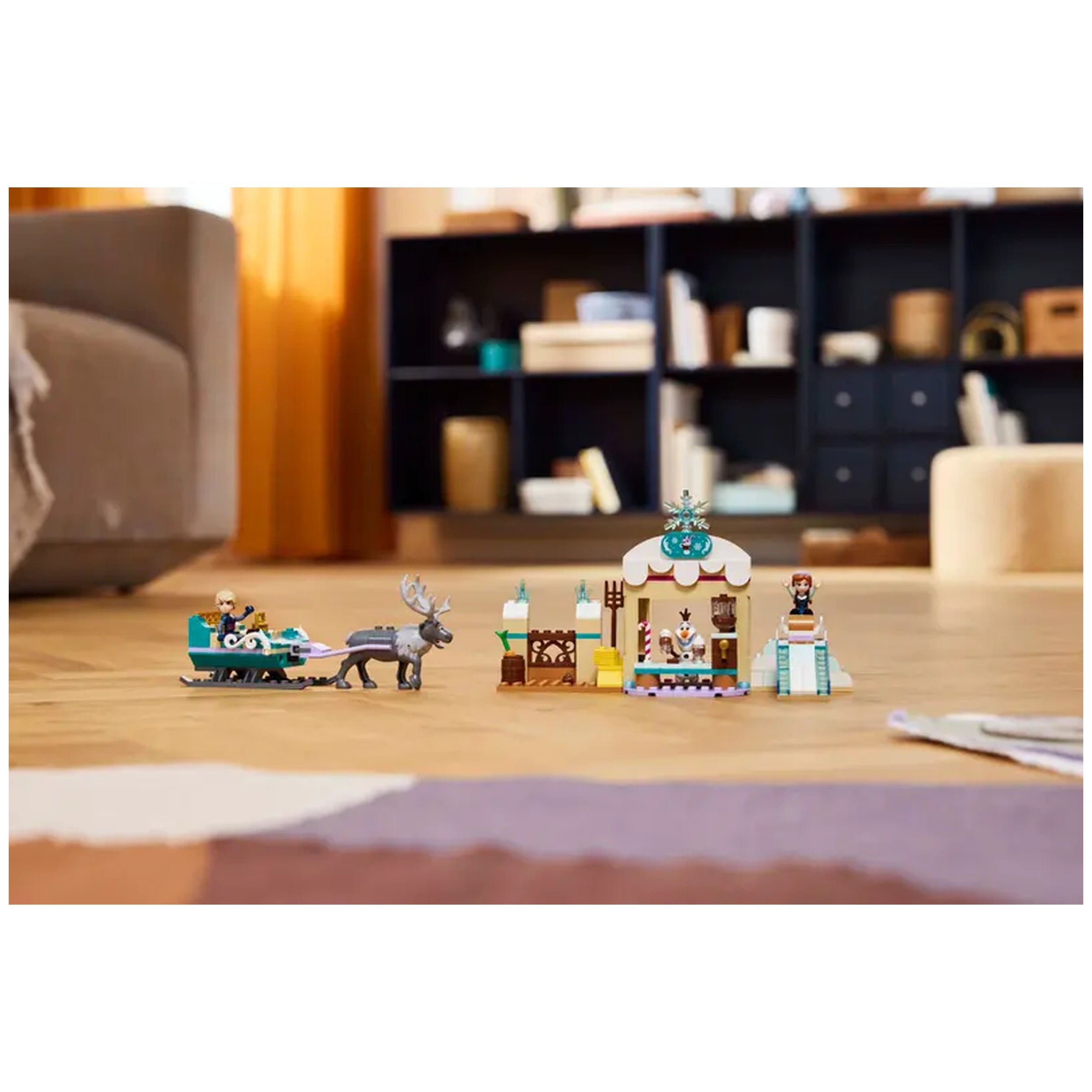 LEGO® Disney's Frozen Anna’s Sleigh Adventure set featuring Anna and Kristoff mini-doll figures, Olaf the snowman, and Sven the reindeer, with a colorful sleigh, hot chocolate stand, and a sledding hill on a wooden floor in a cozy living space.
