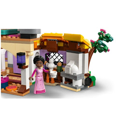A colorful LEGO® construction set depicting Asha's Cottage, featuring a two-level design, intricate details, a character in a pink dress holding two cups, and a cozy kitchen with a decorative table and small accessories. A vibrant tree with pink flowers adds charm to the scene.