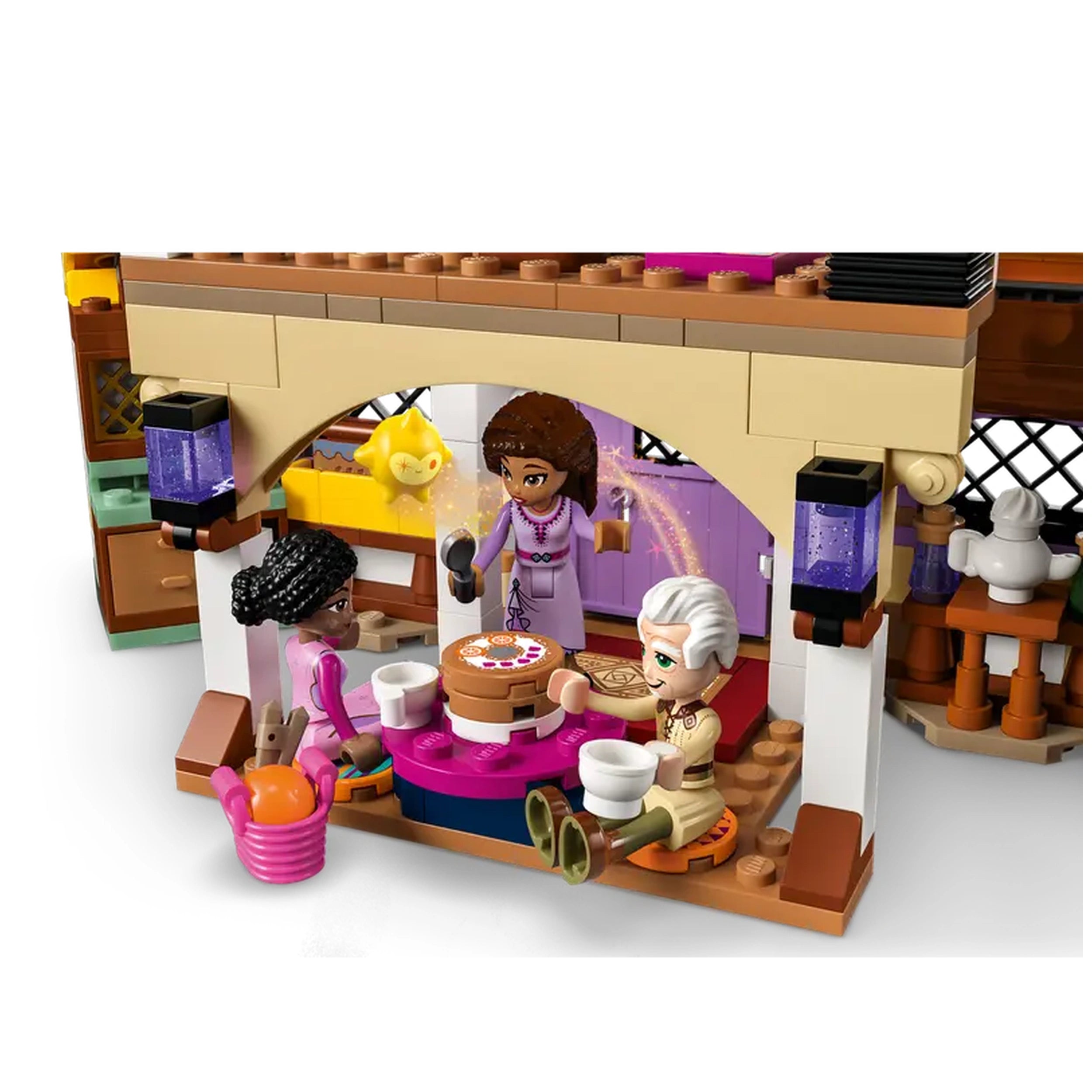 A colorful scene from LEGO® Disney Asha’s Cottage set, featuring three mini dolls enjoying tea at a round table with a cake. The cozy cottage interior showcases a vibrant kitchen and whimsical decor, with a golden character peeking out from above.