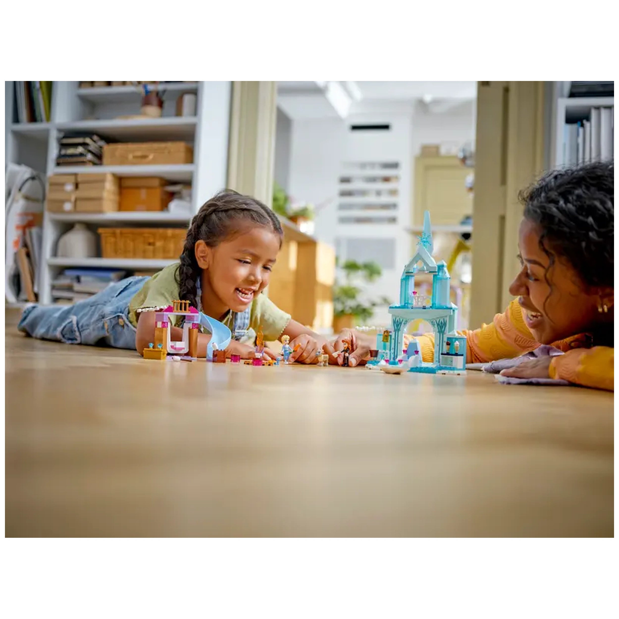 A young girl and an adult woman smile while playing with LEGO Disney Frozen Elsa’s Castle set on a wooden floor, surrounded by colorful building pieces. Brightly lit, the cozy room features shelves and greenery in the background.