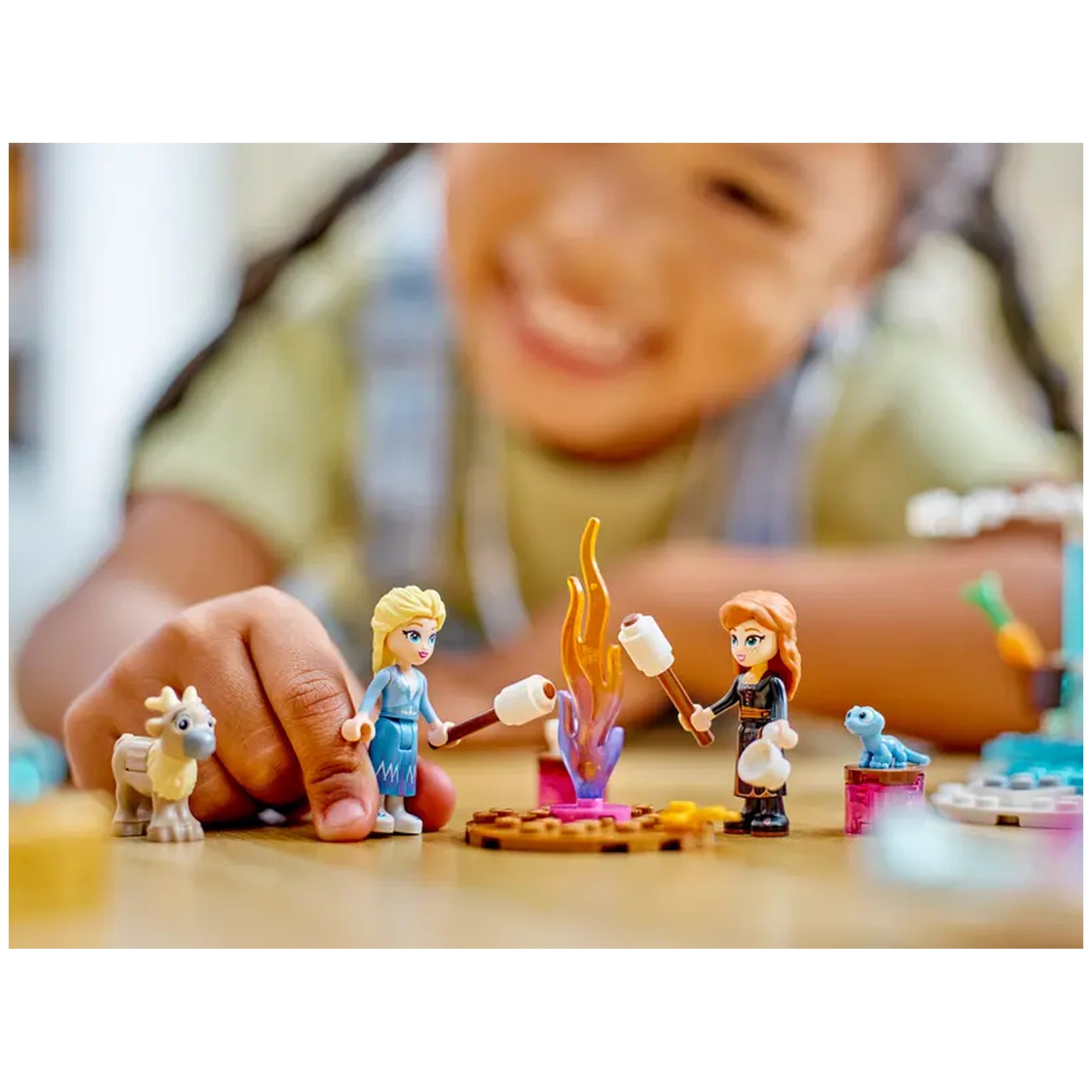 Two LEGO figures of Elsa and Anna roasting marshmallows over a colorful campfire, with a smiling child in the background.