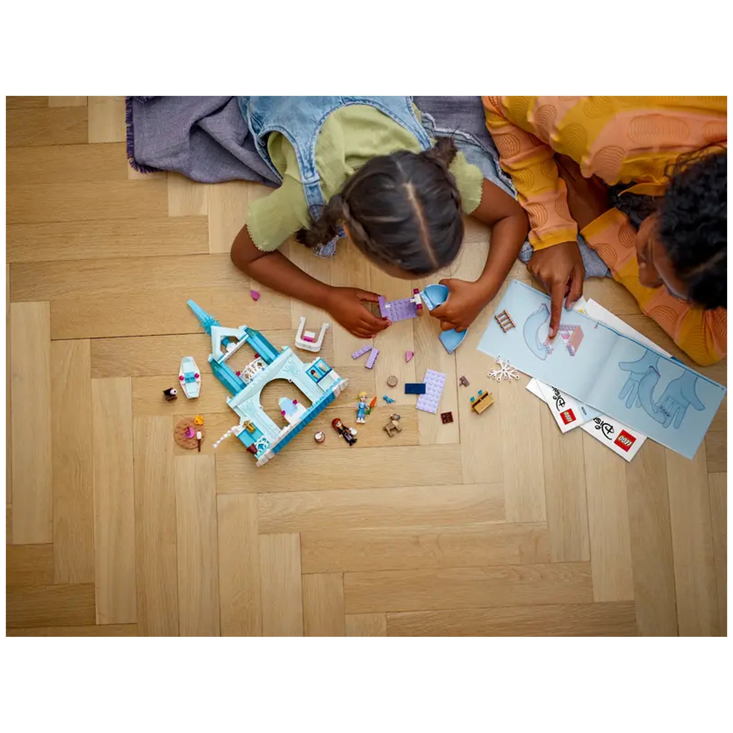 Two children play on a wooden floor, assembling LEGO® Disney Frozen Elsa's castle set. Colorful bricks are scattered around, with an instruction booklet featuring detailed illustrations. One child holds up a purple piece while the other points at the instructions. A cozy, imaginative scene filled with creativity.