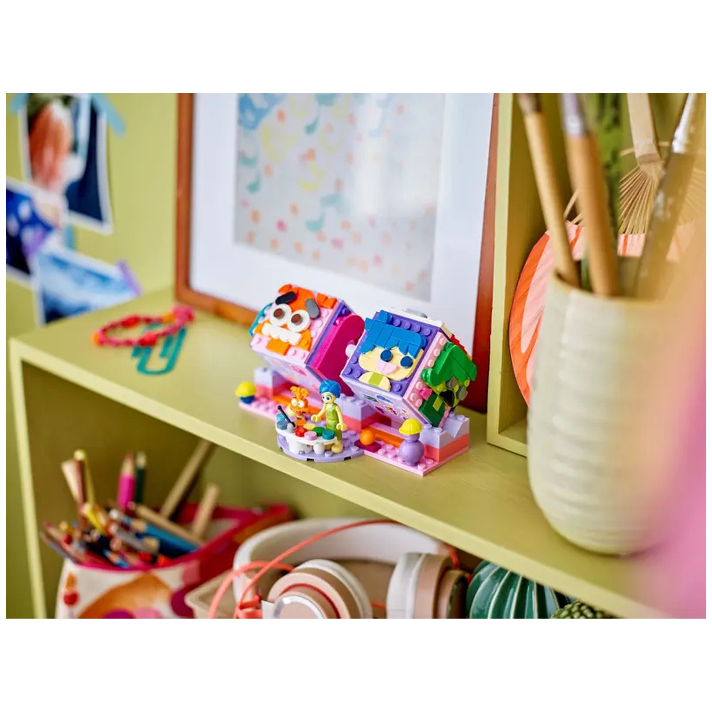 Colorful LEGO Inside Out 2 Mood Cubes featuring Joy and Anxiety figures, displayed on a shelf with art and craft supplies in the background.