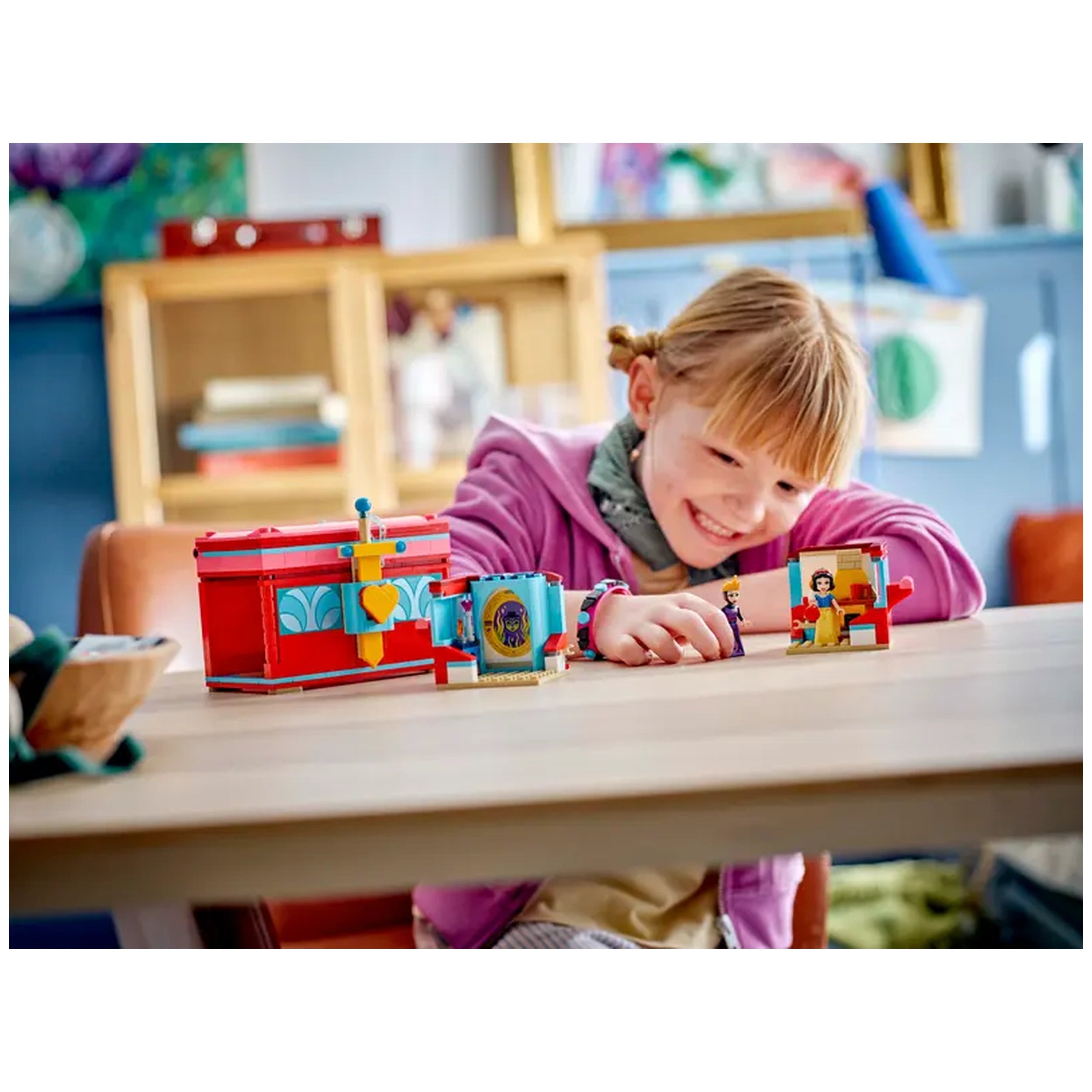 A happy child in a pink jacket plays with a colorful LEGO® Disney Snow White's Jewelry Box set on a wooden table, featuring iconic characters and detailed accessories.