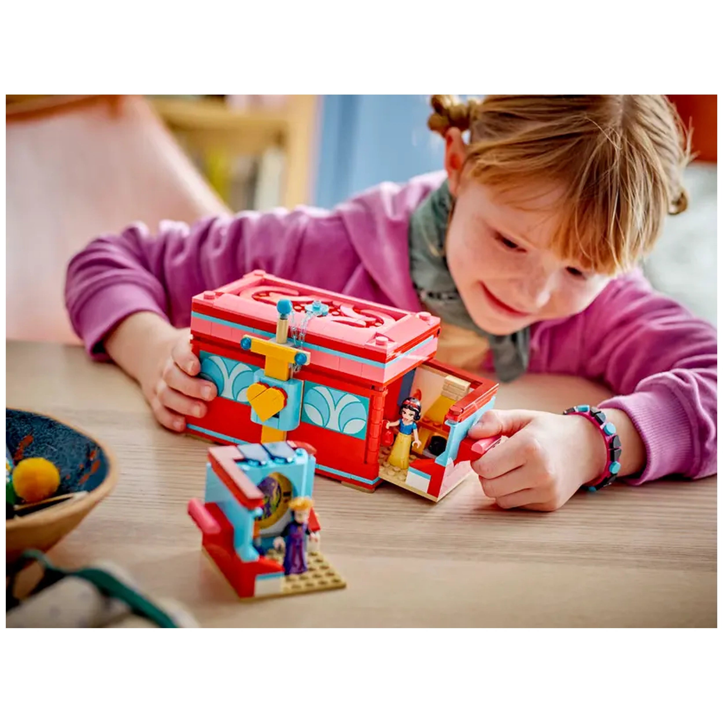 A child wearing a purple hoodie smiles while playing with the red LEGO® Snow White’s Jewelry Box on a wooden table. Two mini-dolls are visible, one of Snow White standing beside the box and another character inside. Colorful detailing and a lockable lid add to the set’s charm.