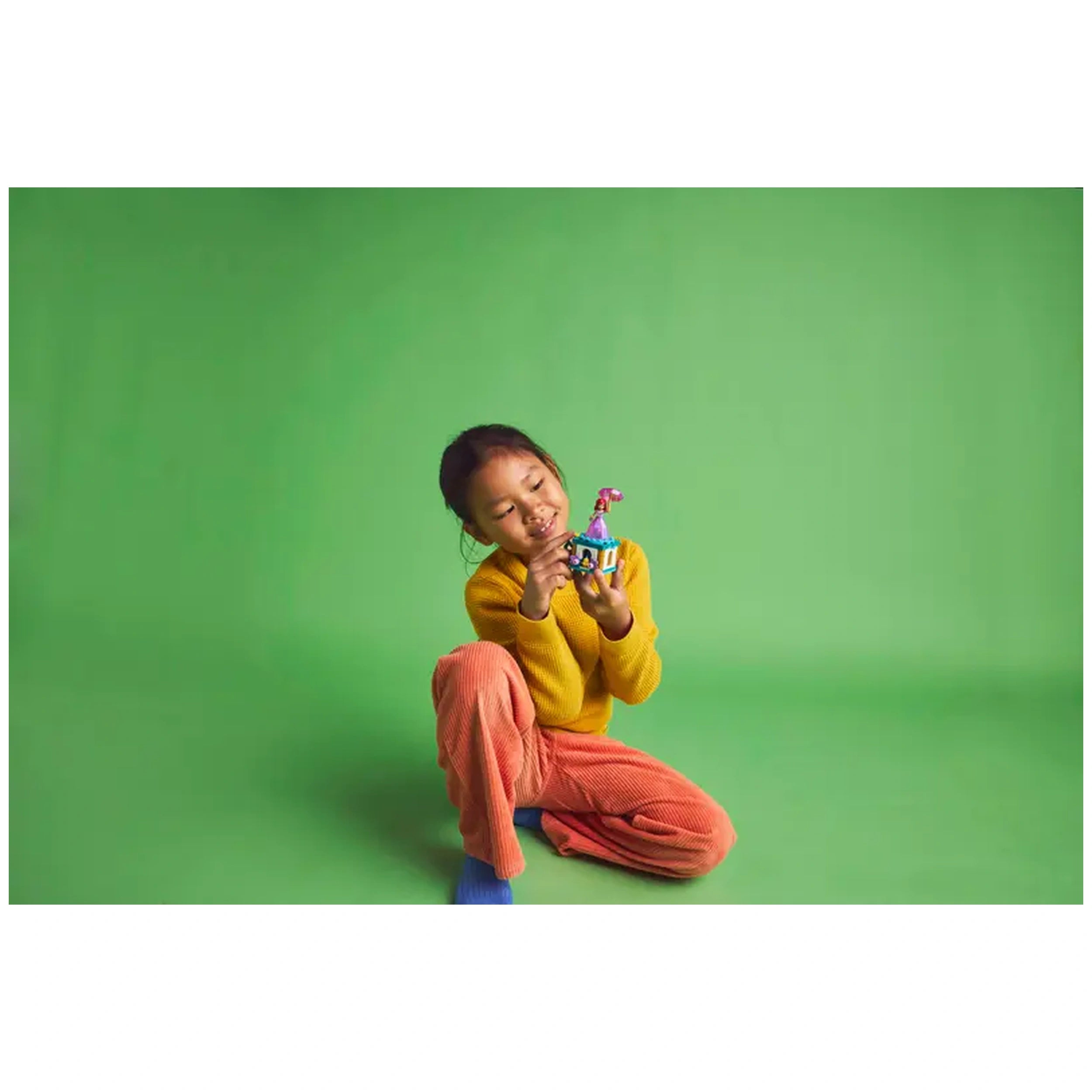 A young girl sits on a green background, smiling while holding the colorful LEGO® Disney Princess Twirling Ariel construction set. She wears a yellow sweater and orange pants, exuding joy as she engages with the toy.