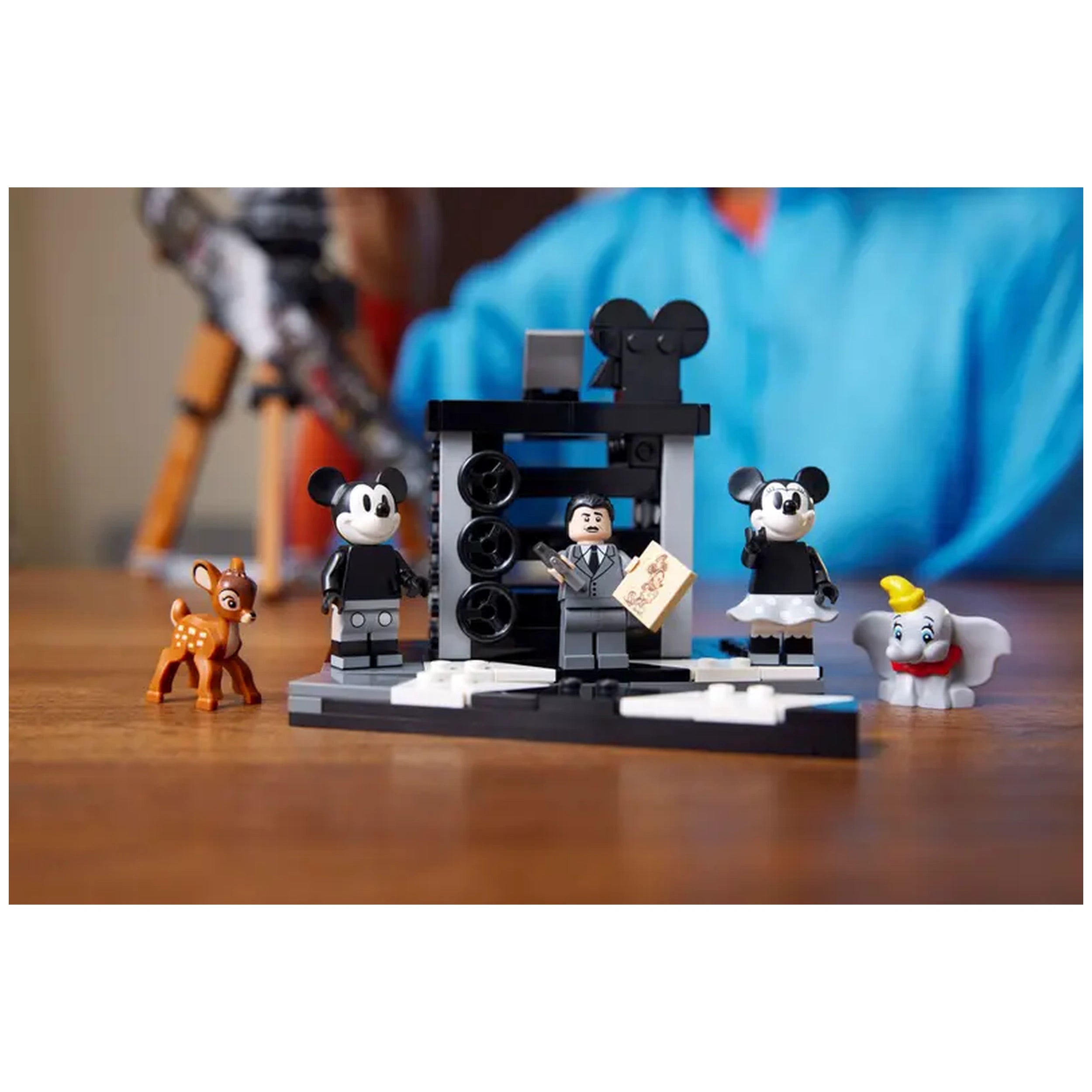 LEGO® Disney Walt Disney Tribute Camera set featuring three minifigures: Mickey Mouse in black attire, a figure resembling Walt Disney holding a script, and Minnie Mouse in a skirt. Two animal figures include Bambi and Dumbo, set against a classic black movie camera backdrop on a wooden surface.