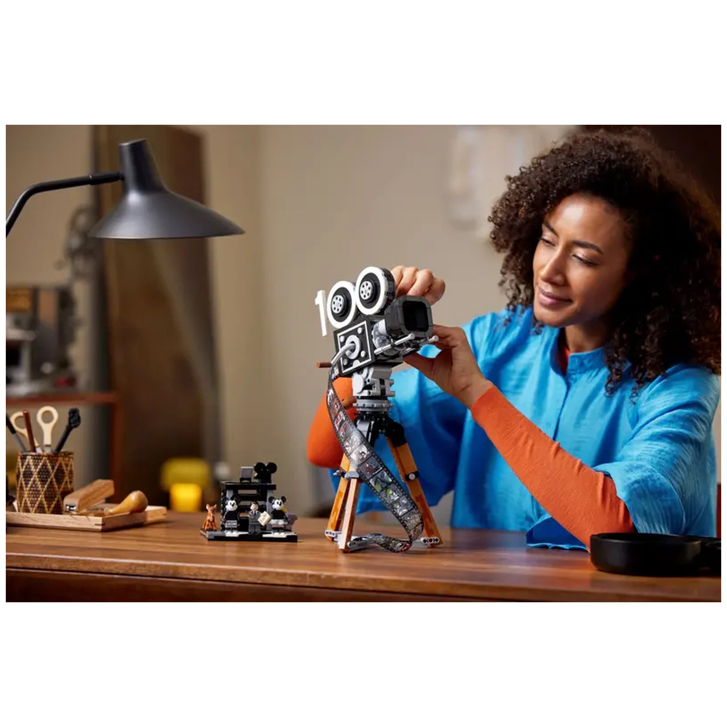 Adult woman in a blue shirt adjusts a LEGO® Disney vintage movie camera on a wooden desk, with a film strip and animal figures nearby.