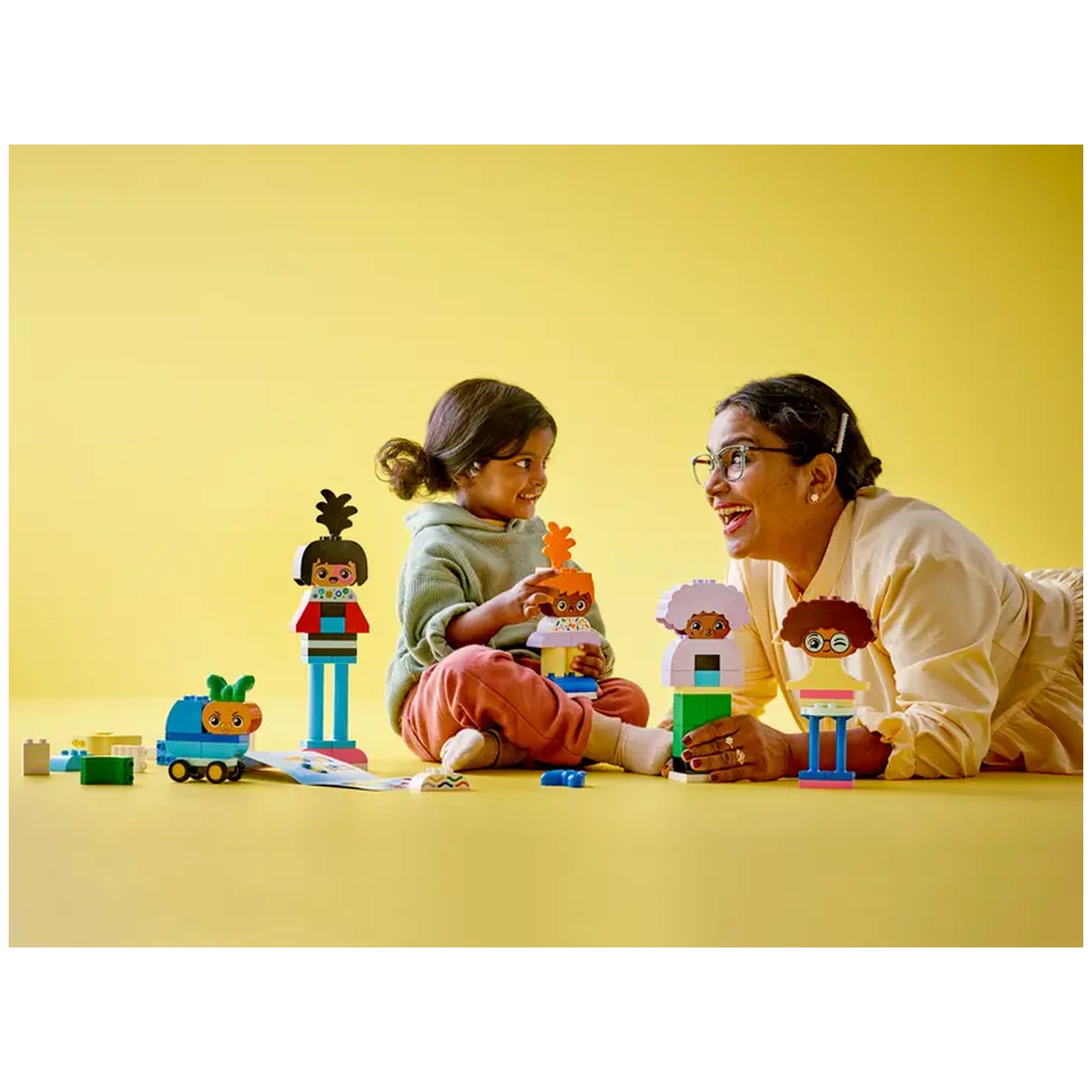A young child and an adult are seated on a yellow floor, joyfully playing with colorful LEGO® DUPLO® figures. The child holds a figure with orange hair and a flower. Several buildable characters and a toy vehicle are visible, demonstrating creative play and emotional exploration. The adult smiles, wearing glasses, as they engage together in a fun learning experience.