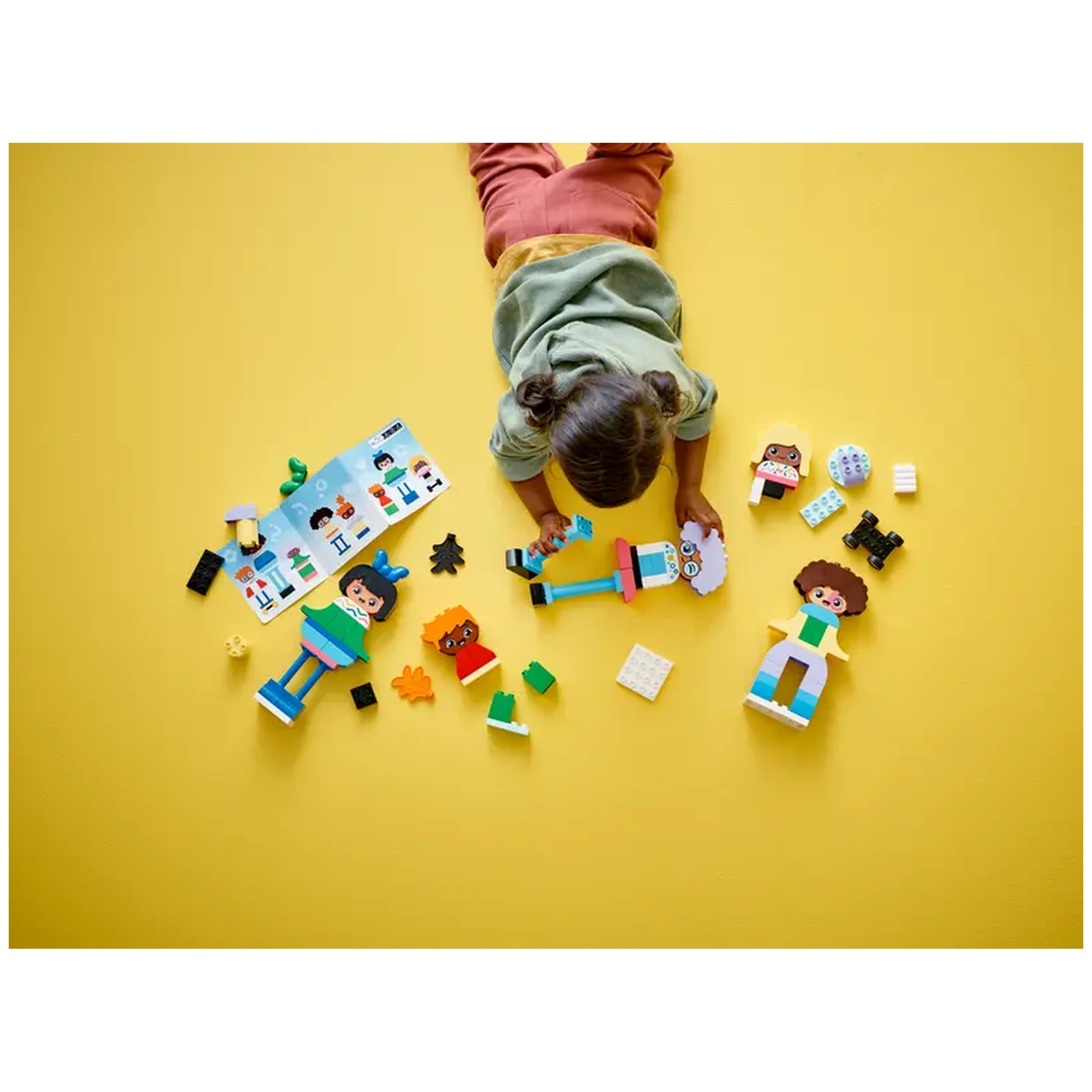 A child lies on a bright yellow floor, playing with LEGO® DUPLO® figures and colorful building blocks. The child is engaged in constructing buildable people, while a user manual and various brick pieces surround them.