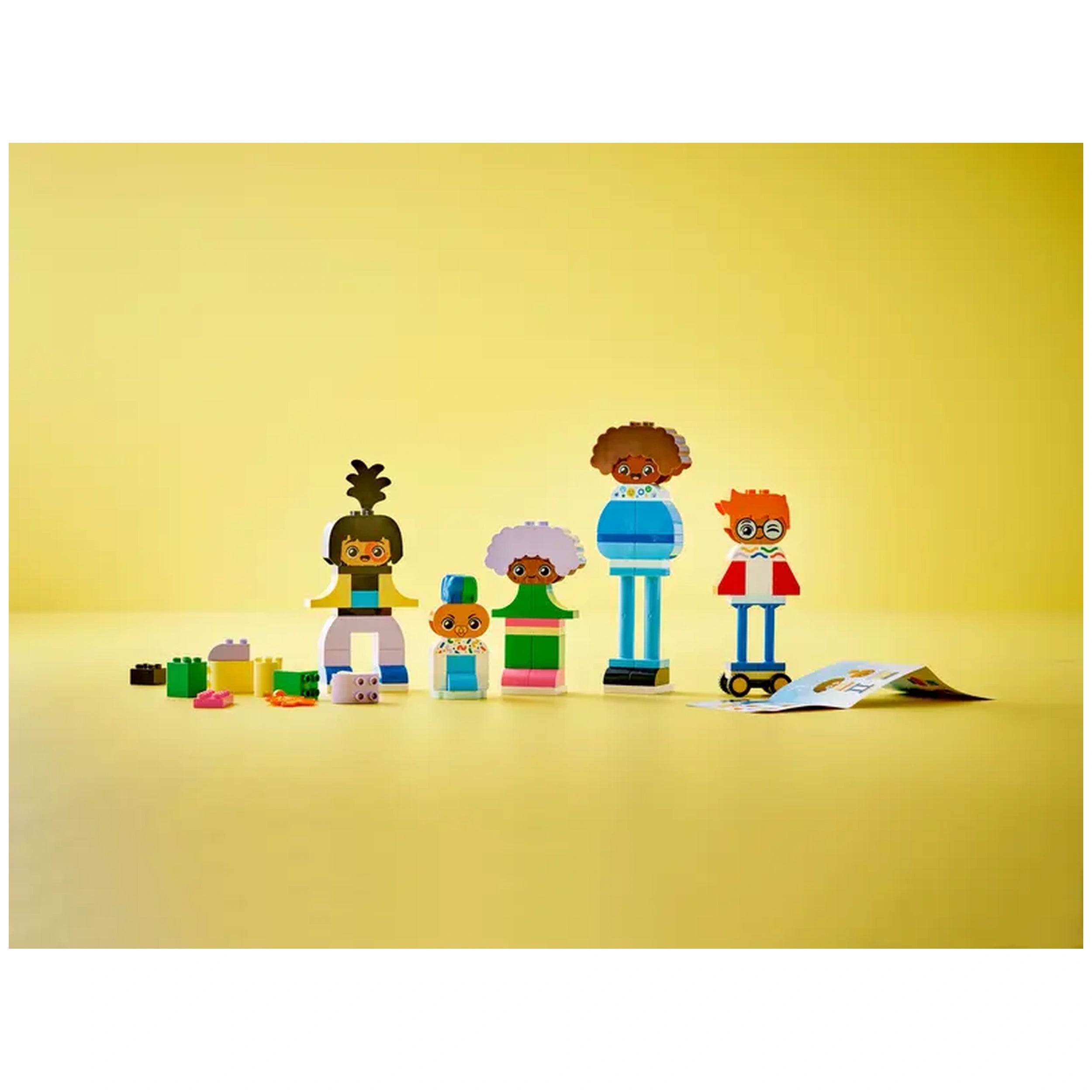 Colorful LEGO® DUPLO® Buildable People characters stand on a yellow background, showcasing a variety of emotions. Alongside, scattered building pieces include bricks and accessories, with an instruction booklet partially visible.