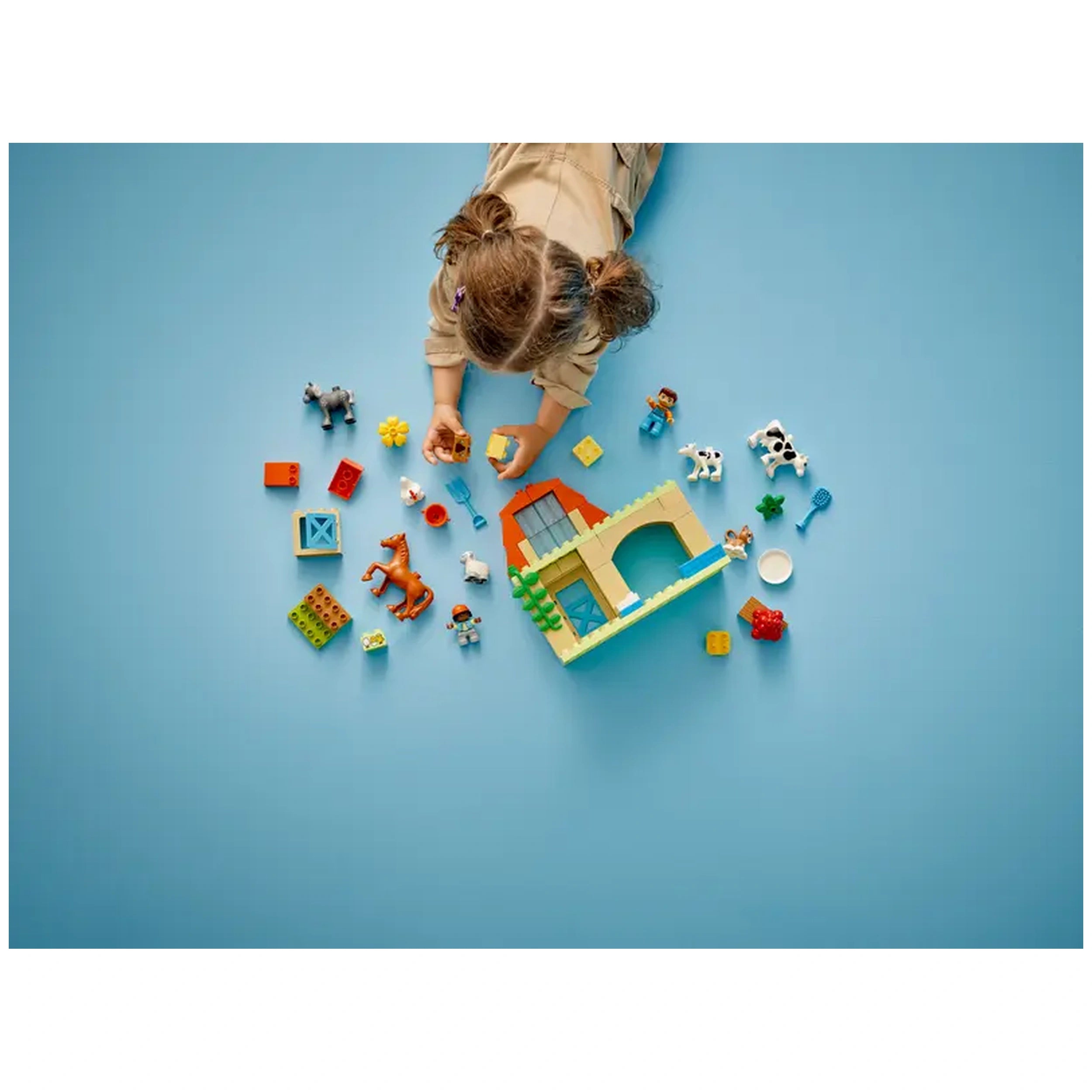 A toddler wearing a beige outfit interacts with colorful LEGO® DUPLO® pieces, including animal figures and a toy farmhouse, on a blue surface.