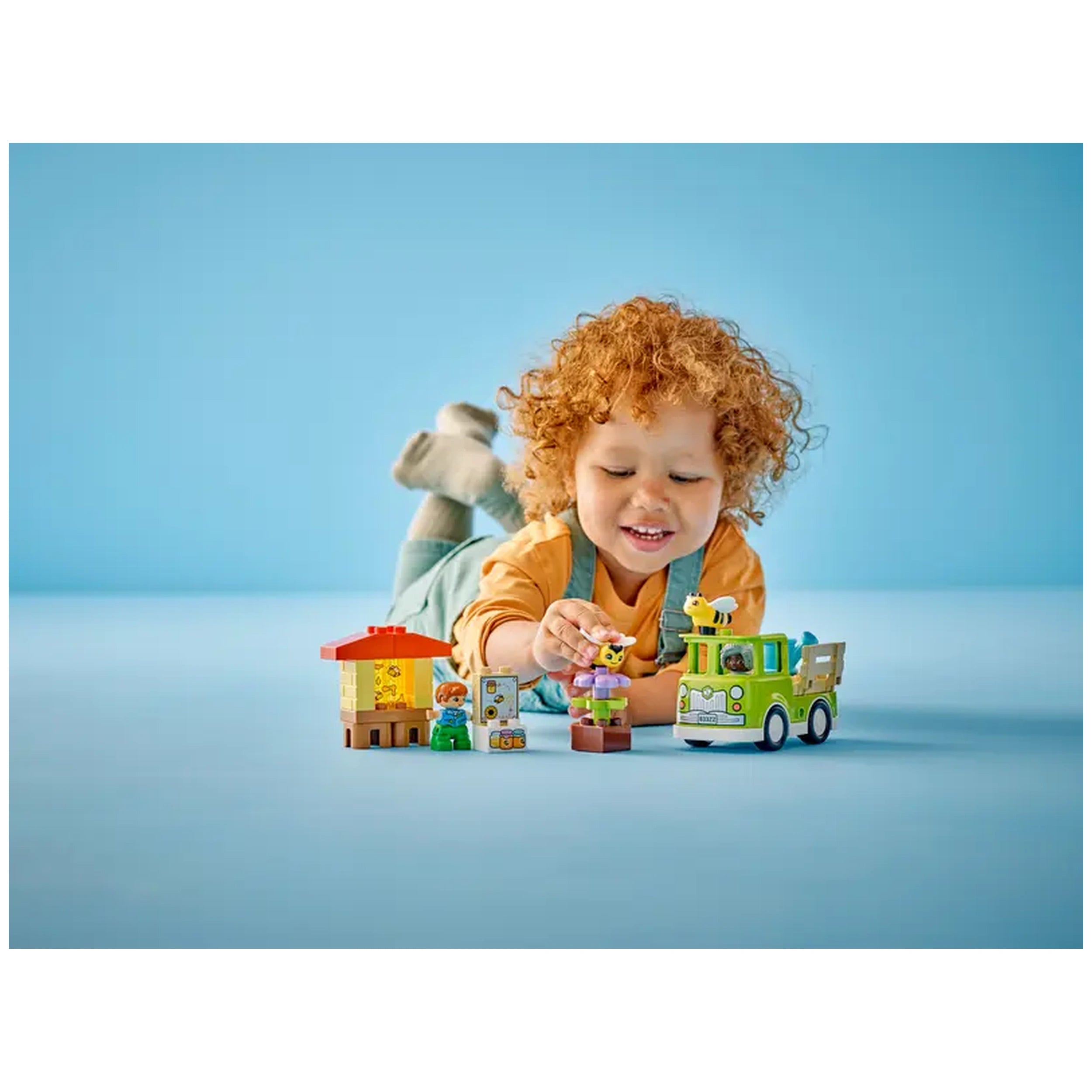 A joyful child with curly red hair plays on the floor, surrounded by a colorful LEGO® DUPLO® set featuring bees, a beehive, and a truck. The child smiles as they hold a bee figure, with vibrant yellow, green, and blue elements in a bright, cheerful setting.