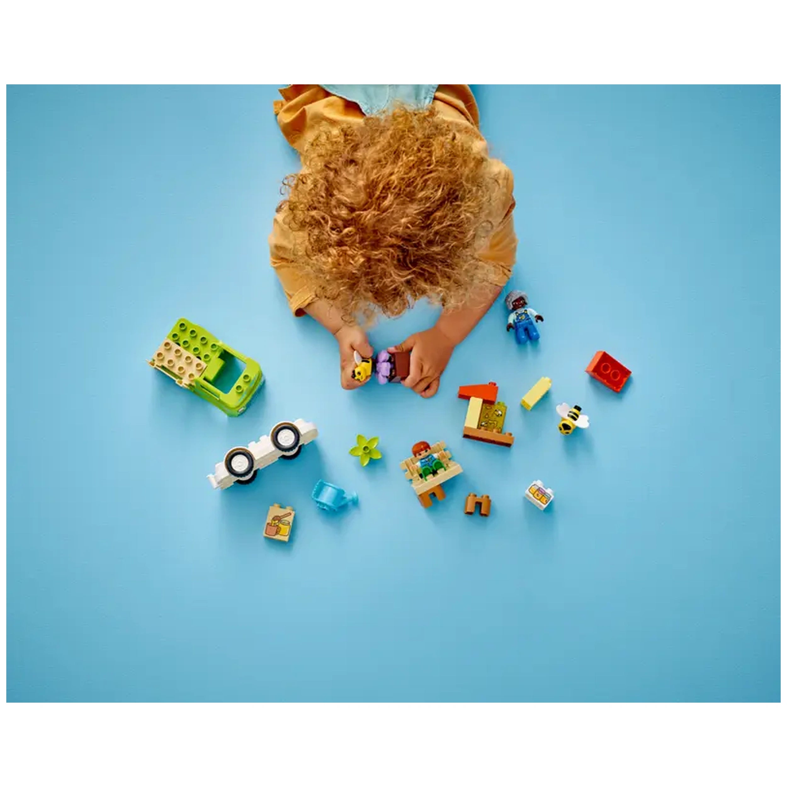 A child with curly hair plays on a blue floor, engaging with colorful Lego pieces, including a truck, bee figurines, and various flower elements.